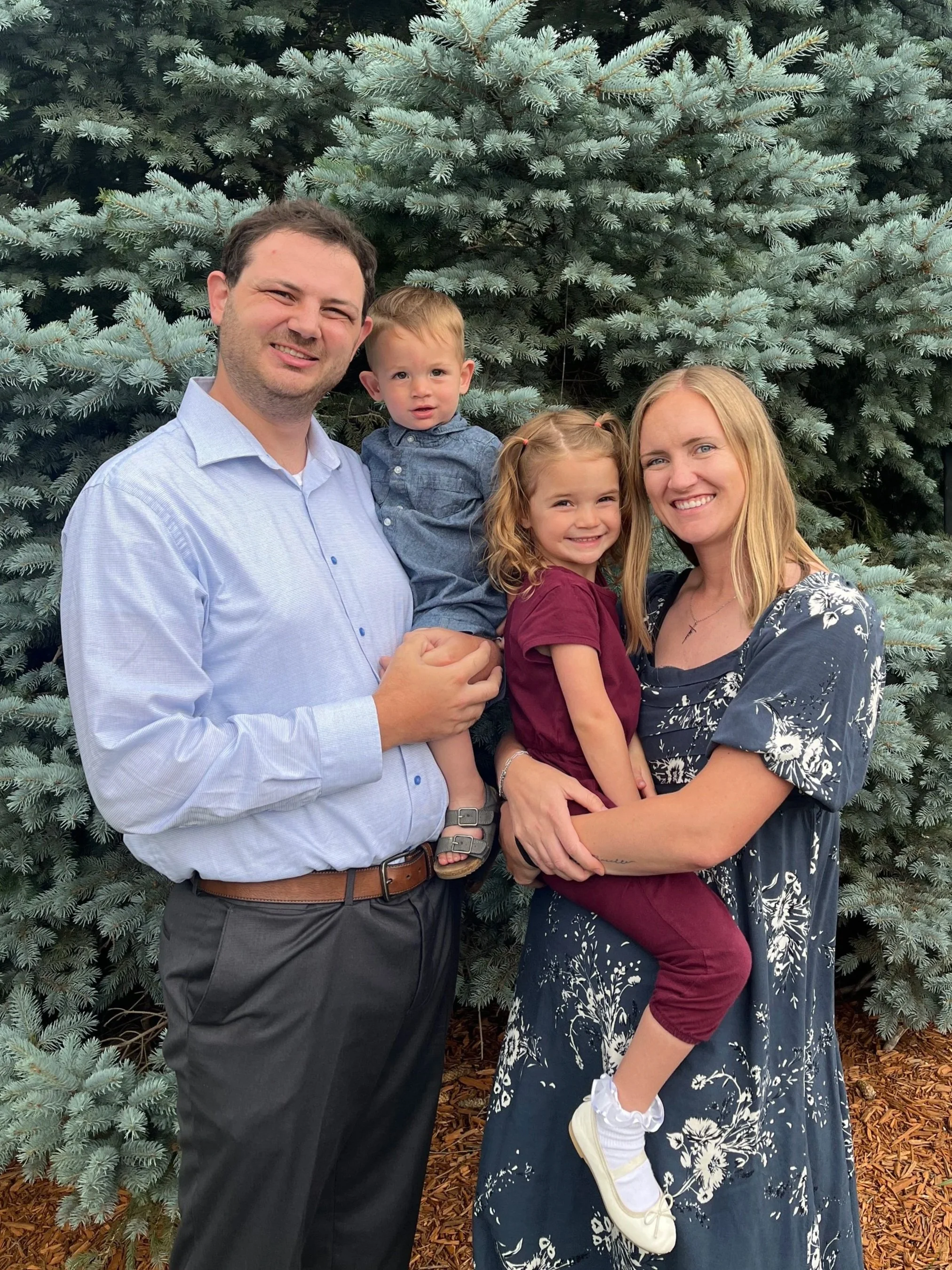 Family of four standing in front of a large green pine tree outdoors. The father is wearing a light blue dress shirt, black pants, and a belt. The mother is wearing a navy blue dress with white floral patterns. The son, who is being held by his father, has light brown hair and is wearing a denim shirt. The daughter, held by her mother, has blonde hair tied in pigtails and is wearing a maroon shirt, maroon pants, white sneakers, and white socks with frills.