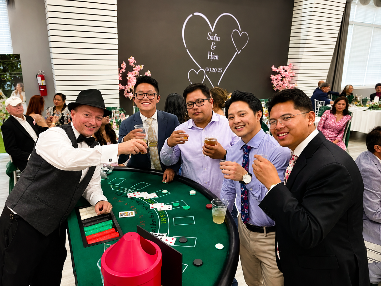 A group of five men gathered around a craps table at a celebration, raising drinks and smiling, with other guests in the background and pink floral decorations behind them.