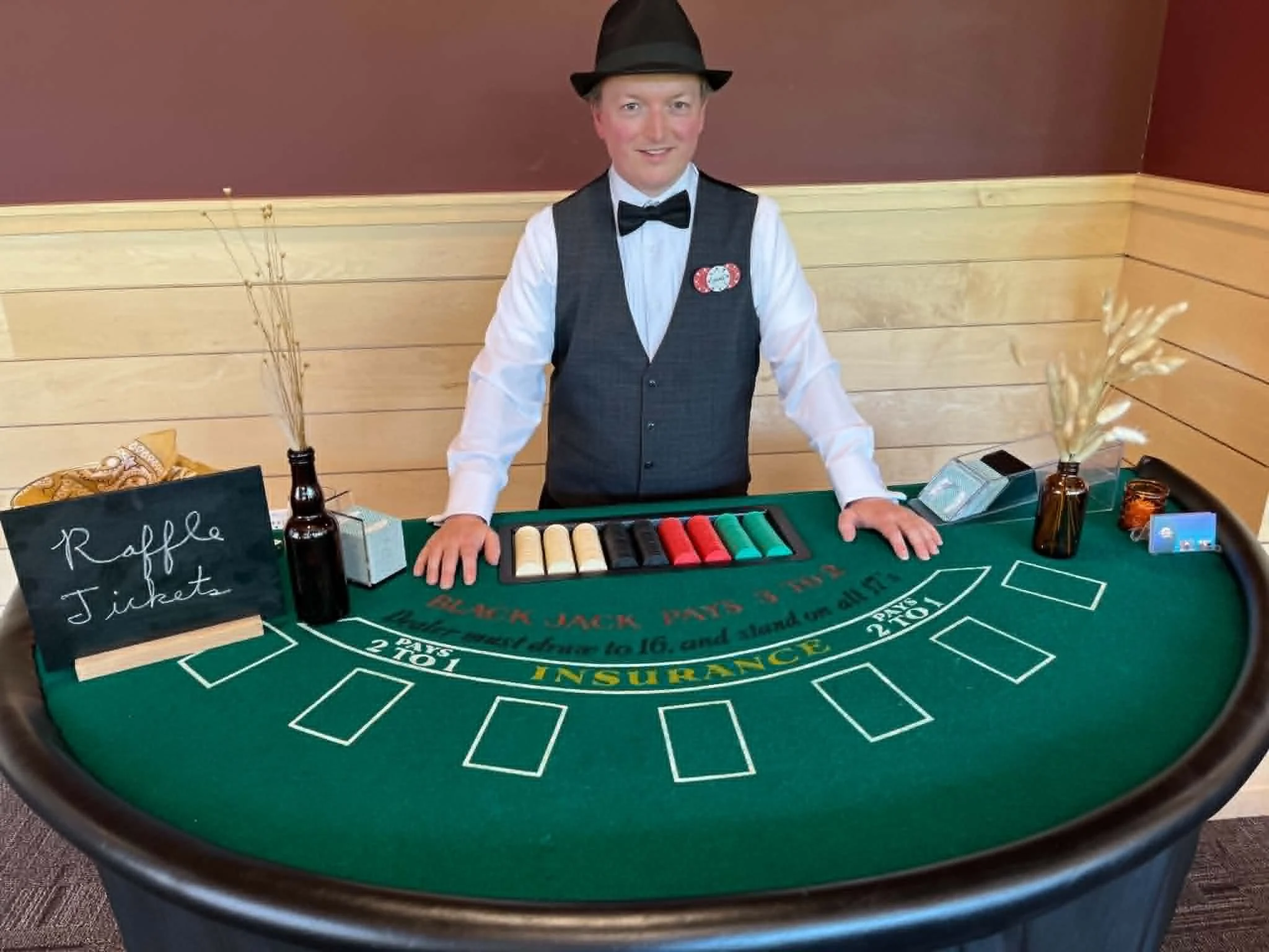 A young man dressed as a dealer in casino attire standing behind a blackjack table. He is wearing a black hat, white shirt, black vest, and a bow tie. The table has a green felt surface with betting areas and chips arranged on it. There are decorative plants and bottles on the table, and a blackboard sign that says 'Raffle Tickets.' The background features a wooden wall with a reddish-brown paint.