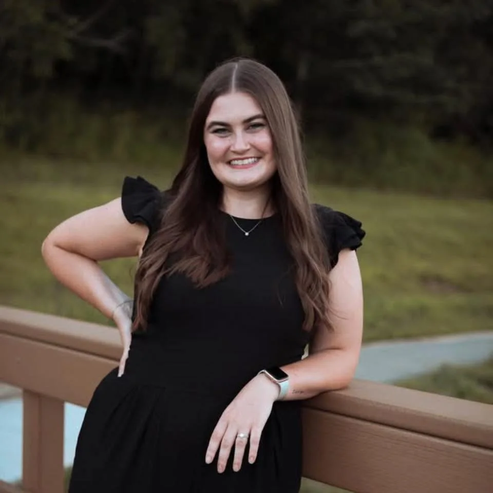A smiling woman with long brown hair wearing a black dress and a smartwatch on her left wrist, standing outdoors with a wooden railing and greenery in the background.
