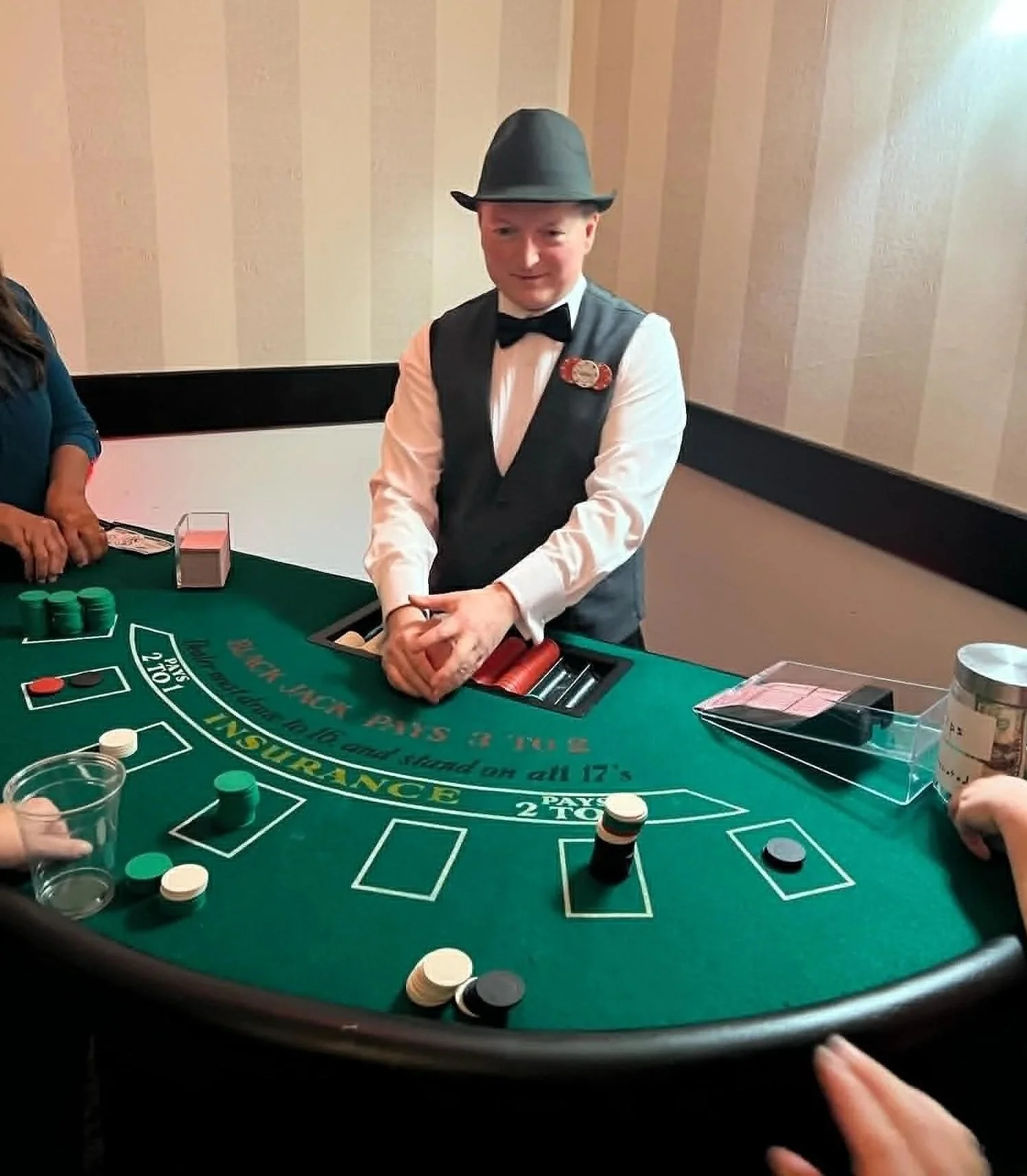 A young boy dressed in a tuxedo vest, white shirt, bow tie, and a fedora hat, standing at a craps table in a casino. He appears to be rolling dice while other players' hands are visible around the table.