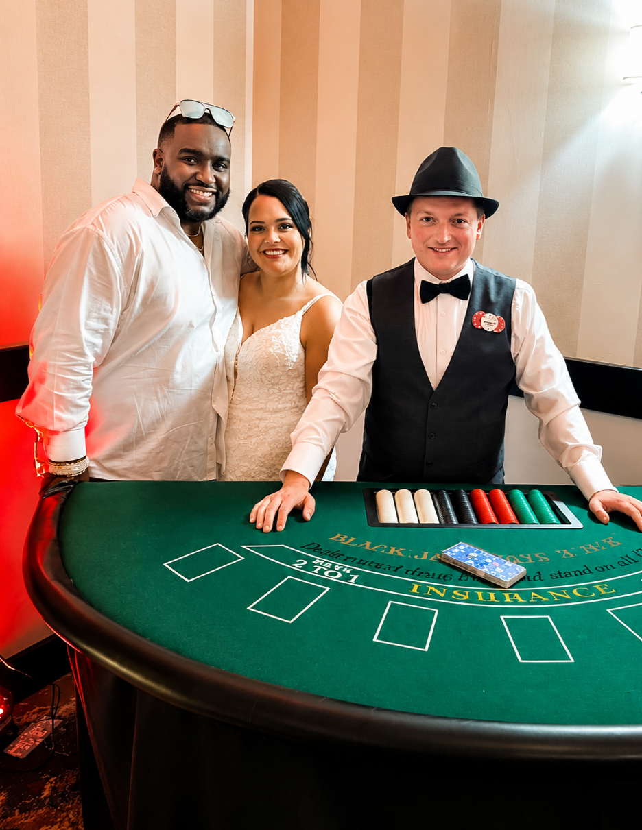 Three people standing behind a casino blackjack table, two men and one woman, smiling for the camera. The man on the right is dressed as a dealer with a black vest, bow tie, and hat. The woman is in a white dress, and the man on the left is wearing glasses and a white shirt. The table has chips and a deck of cards on it.