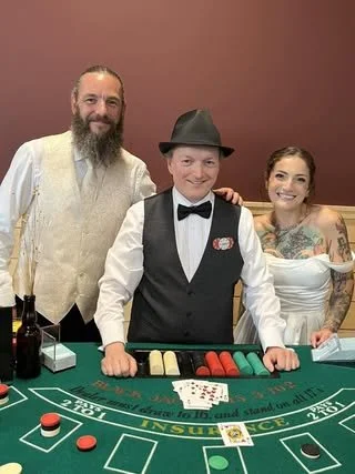 A dealer in a tuxedo and hat standing behind a roulette table with a smiling woman wearing a white strapless dress. The woman has tattoos on her arms and a big smile.