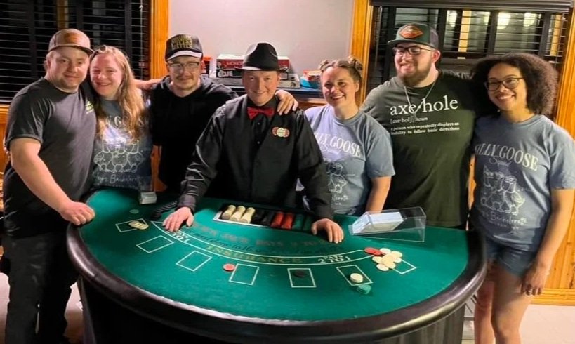 Group of six people standing around a blackjack table, smiling, with a dealer in the center wearing a black shirt, red bow tie, and a fedora hat. Some people are wearing casual t-shirts, one with a 'Bully Goose' logo, and they are inside a room with wood-paneled walls and large windows.