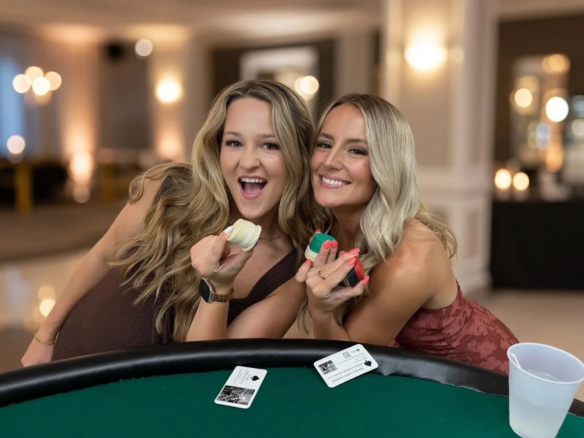 Two women smiling and holding macarons, sitting at a gaming table with poker chips and playing cards in a decorated room.