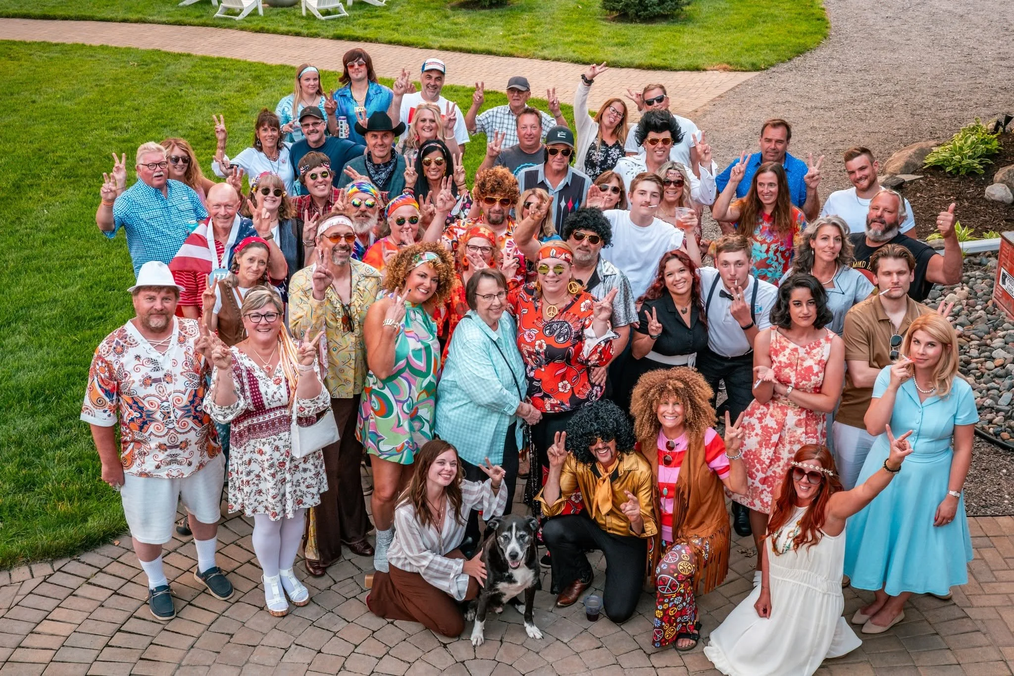 A large group of people dressed in vibrant, vintage, and hippie-inspired clothing posing outdoors on a paved area with grass and trees in the background. Many are making peace signs and smiling, with a black and white dog in front.