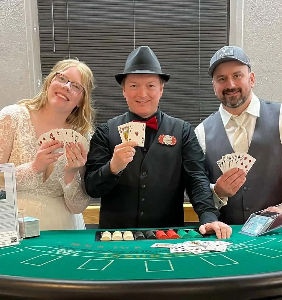 Three people dressed in formal and vintage clothing posing with playing cards and poker chips at a casino-style table.