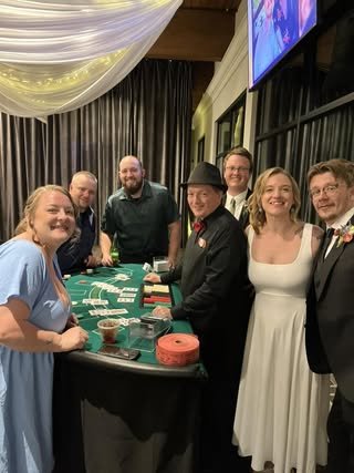 Group of seven people dressed in formal attire gathered around a blackjack table in a casino-themed setting, celebrating an event.