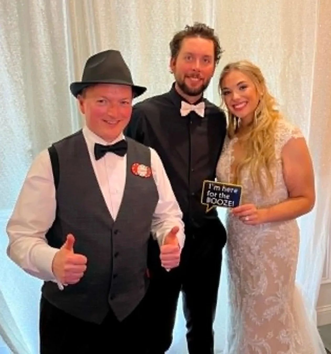 Three people dressed in formal attire at a celebration. One man in a vest, bow tie, and hat giving thumbs up, another man in a black suit and bow tie, and a woman in a lace wedding gown holding a sign that says "Top of the GROC". They are smiling and standing against a light curtain background.