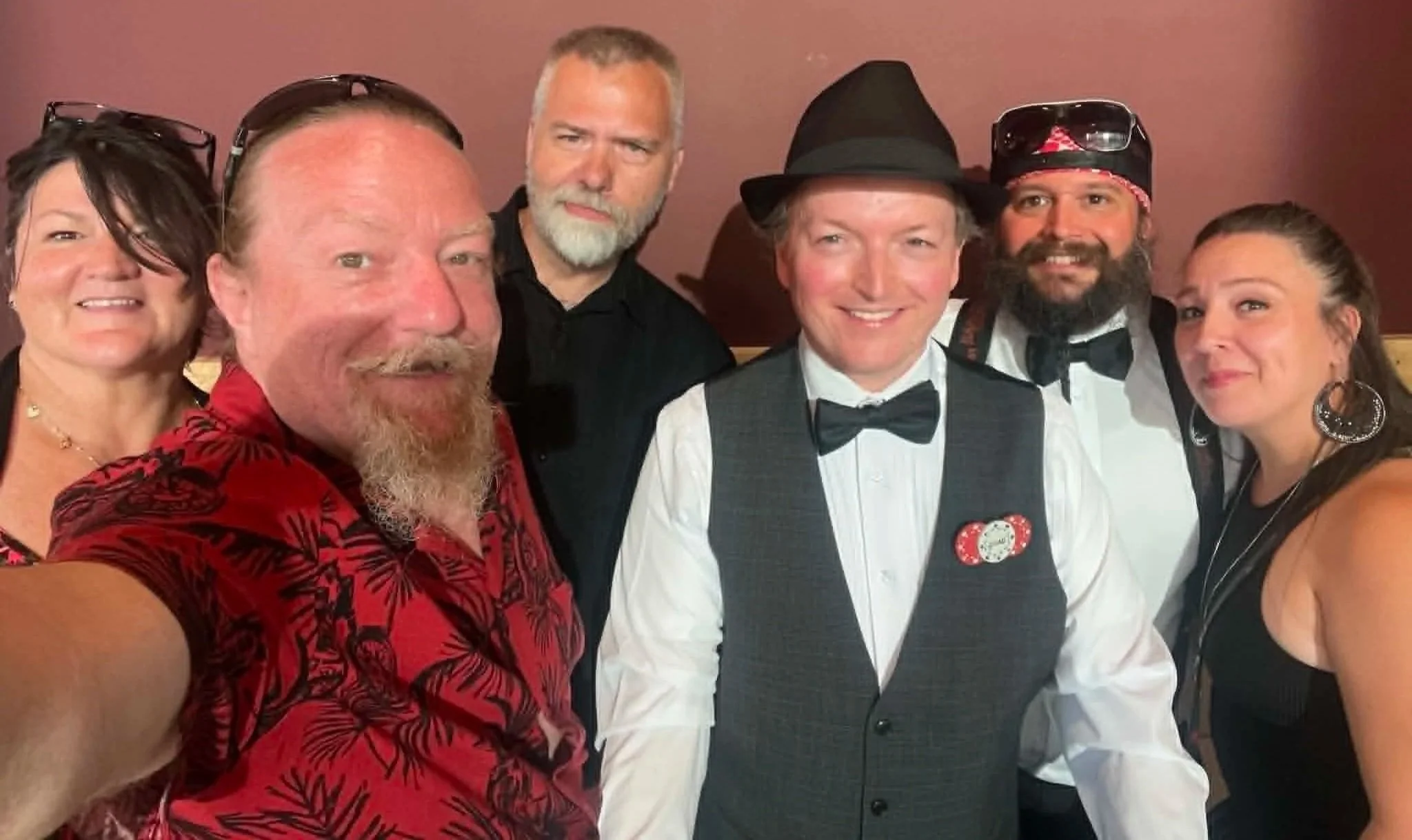 Group of seven people smiling and posing for a selfie at a party, dressed in semi-formal attire with some wearing hats and accessories, standing against a plain brown wall.