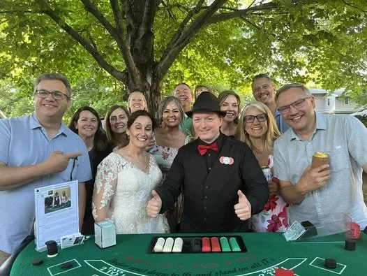 Group of smiling people posing around a casino blackjack table outdoors under a tree, with a dealer in black clothing and a hat in the center.