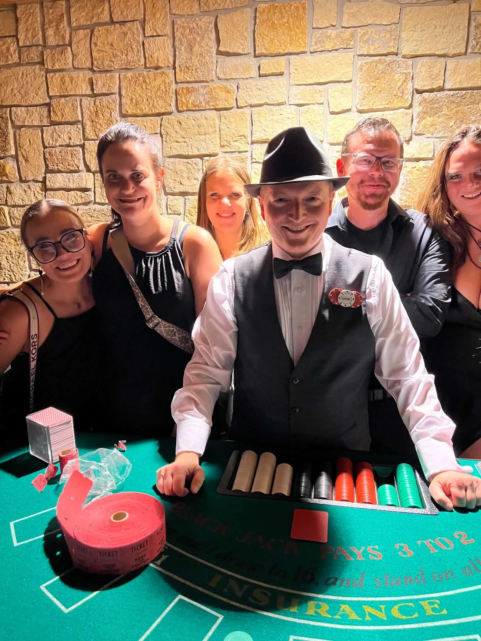 Group of six people gathered around a casino blackjack table, smiling and posing for the photo. One man in the center wears a black hat, bow tie, and vest. The table features poker chips, a deck of cards, and a red tape roll labeled 'TICKET' in front. The background is a wall made of beige bricks.