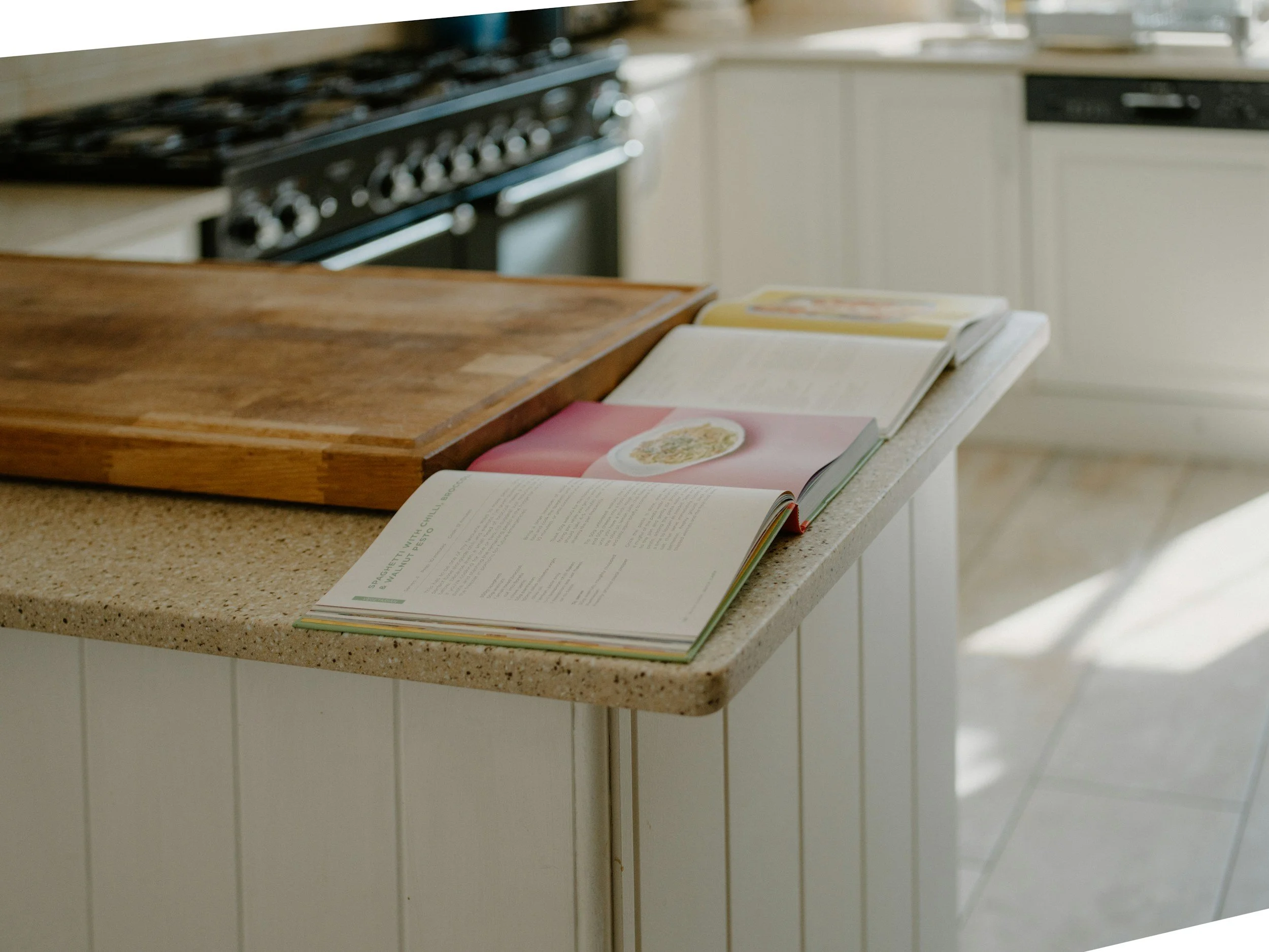 Open cookbooks spread out on kitchen counter with wooden cutting board nearby, in a bright kitchen setting.