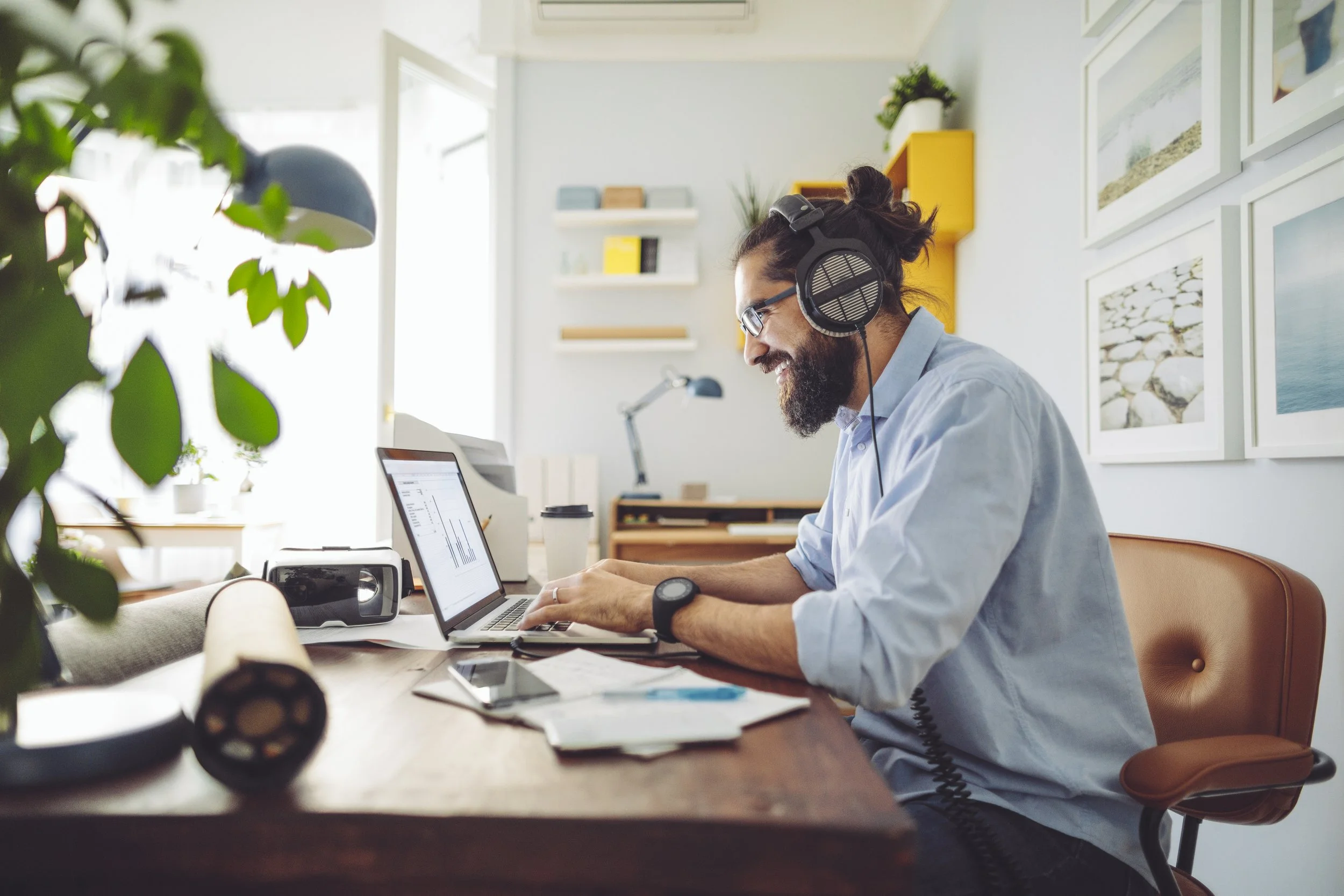 A man with glasses and a beard wearing headphones and a light blue shirt sitting at a desk working on a laptop, with various papers, a smartphone, and office equipment on the desk, in a bright and modern office.