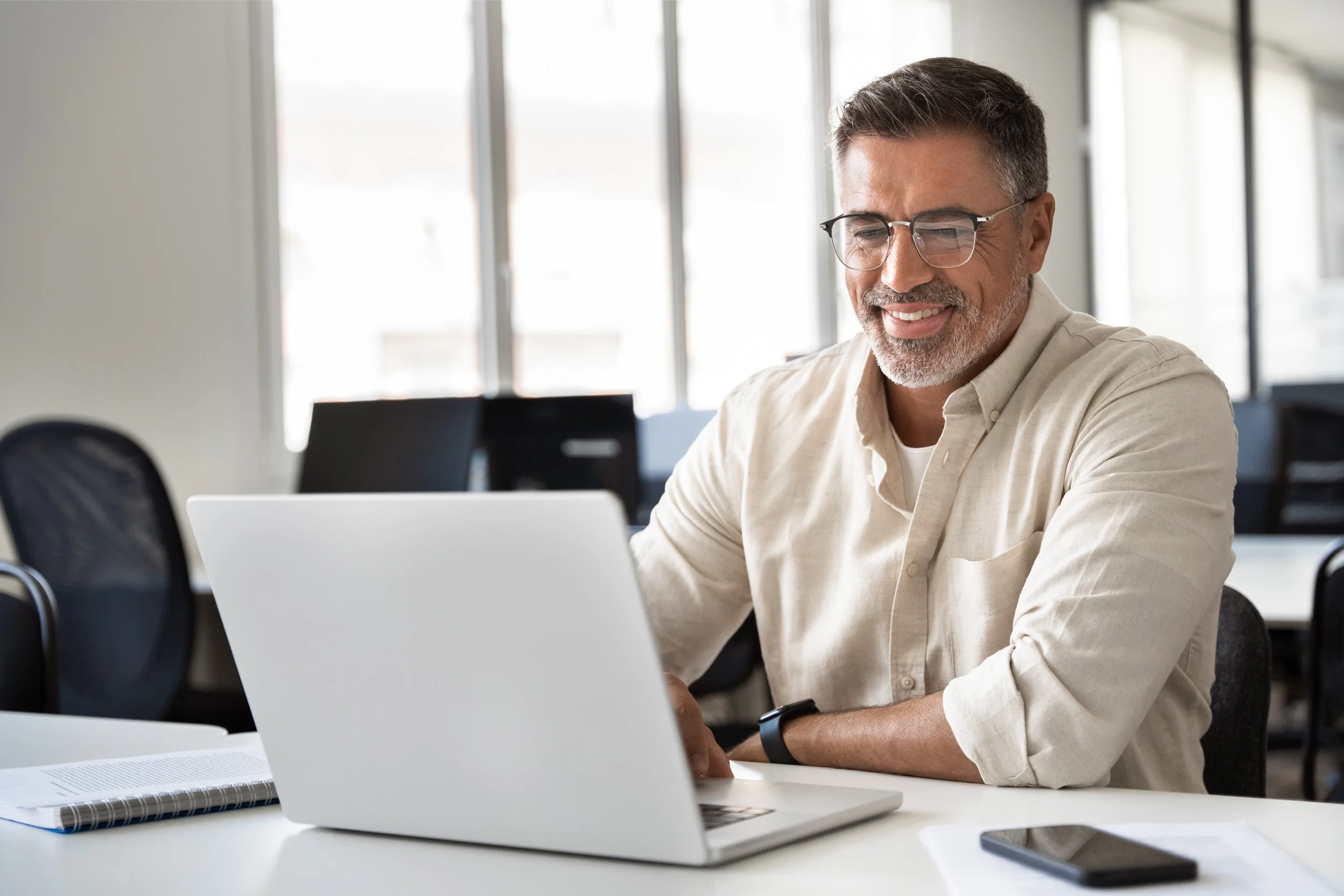 A middle-aged man with gray hair, glasses, and a beard, sitting at a desk in a bright office, smiling while working on a laptop.