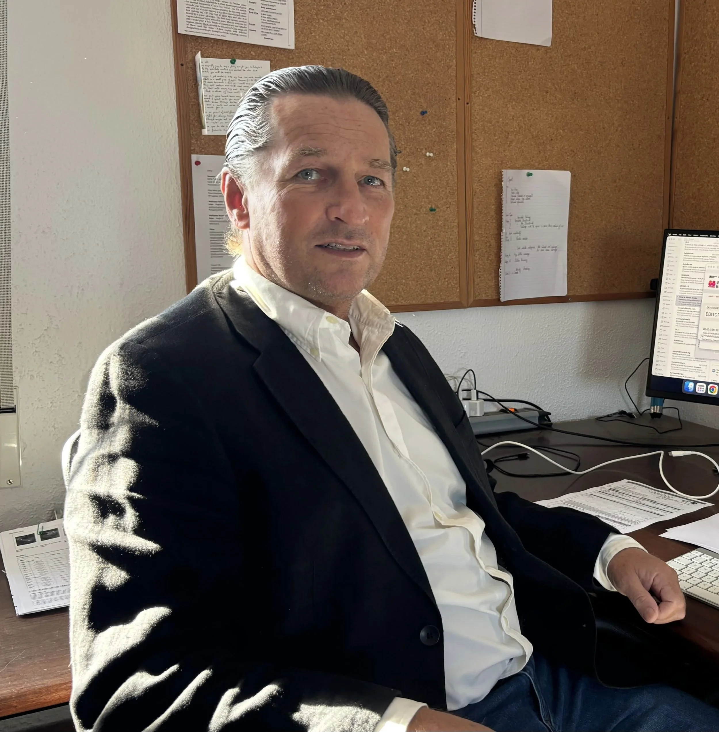 A man with slicked-back hair and blue eyes, wearing a white shirt and dark blazer, sitting at a desk in an office environment. Behind him is a corkboard with papers pinned on it, and on his desk, there are papers, a computer monitor, and a keyboard.