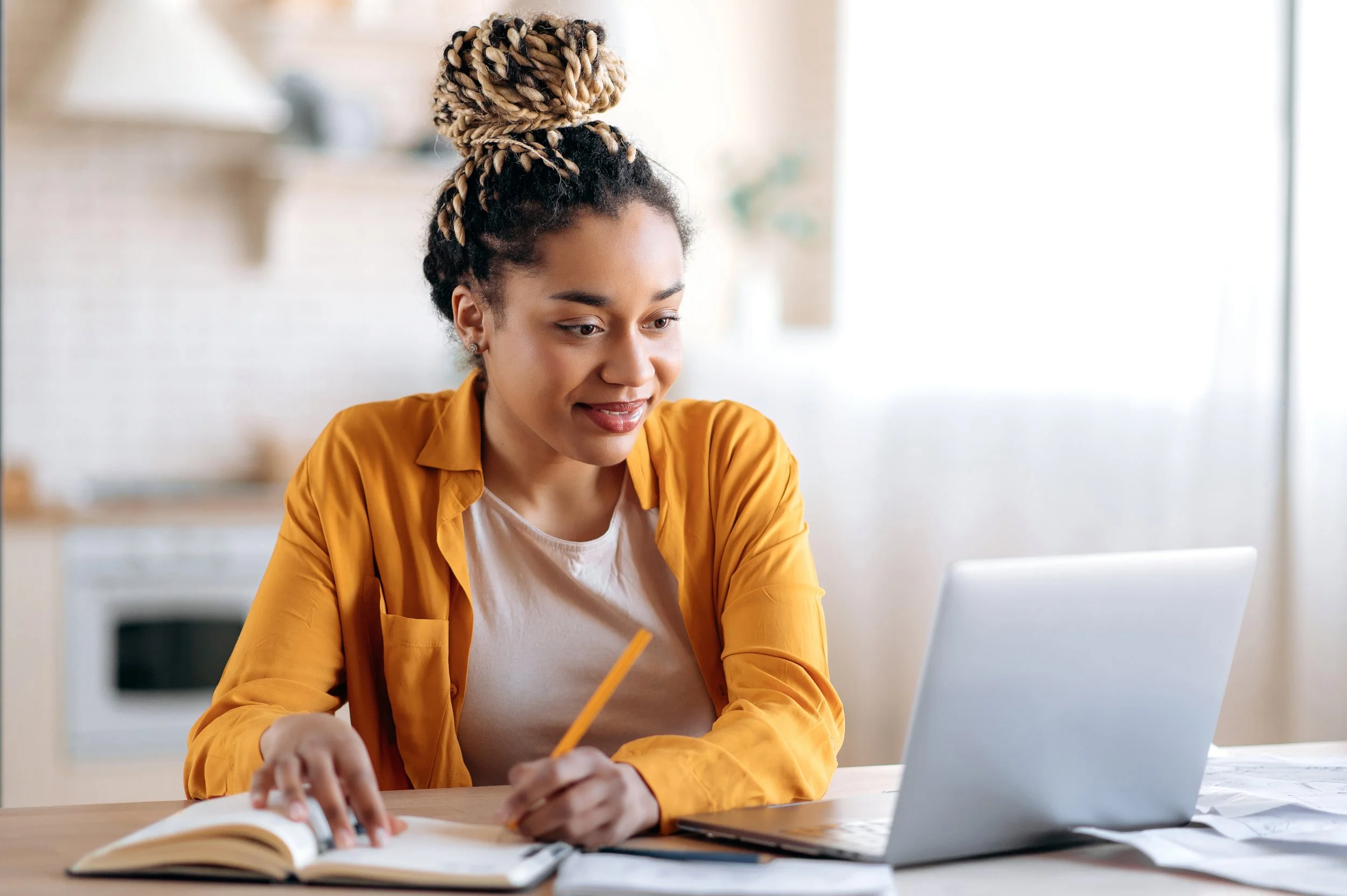 A woman with curly hair styled in a high bun with blonde extensions, working at a desk with a laptop, notebook, and pencil, in a bright, modern kitchen.