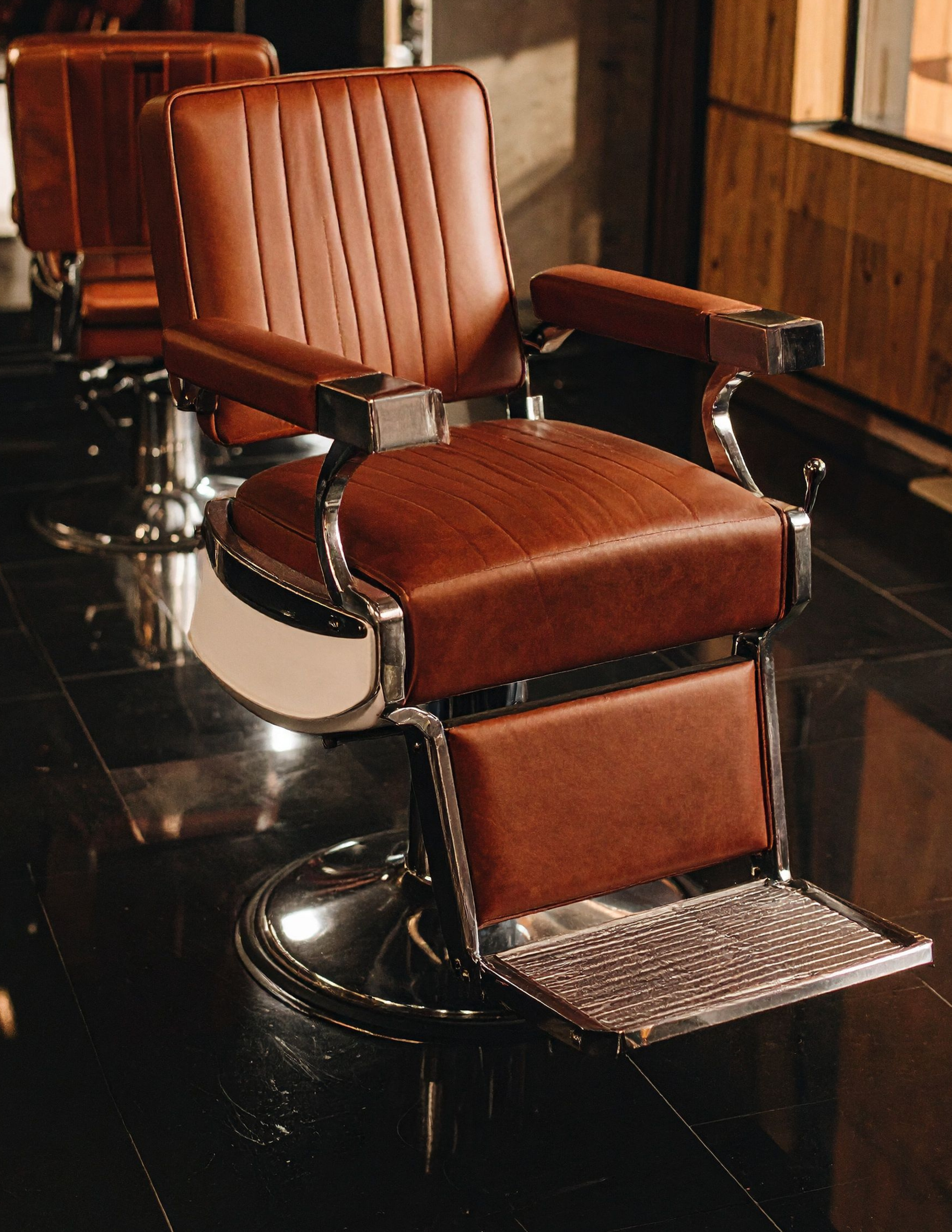 Brown vintage barber chair with metal frame in a dimly lit barbershop with black tiled floor and wooden wall.