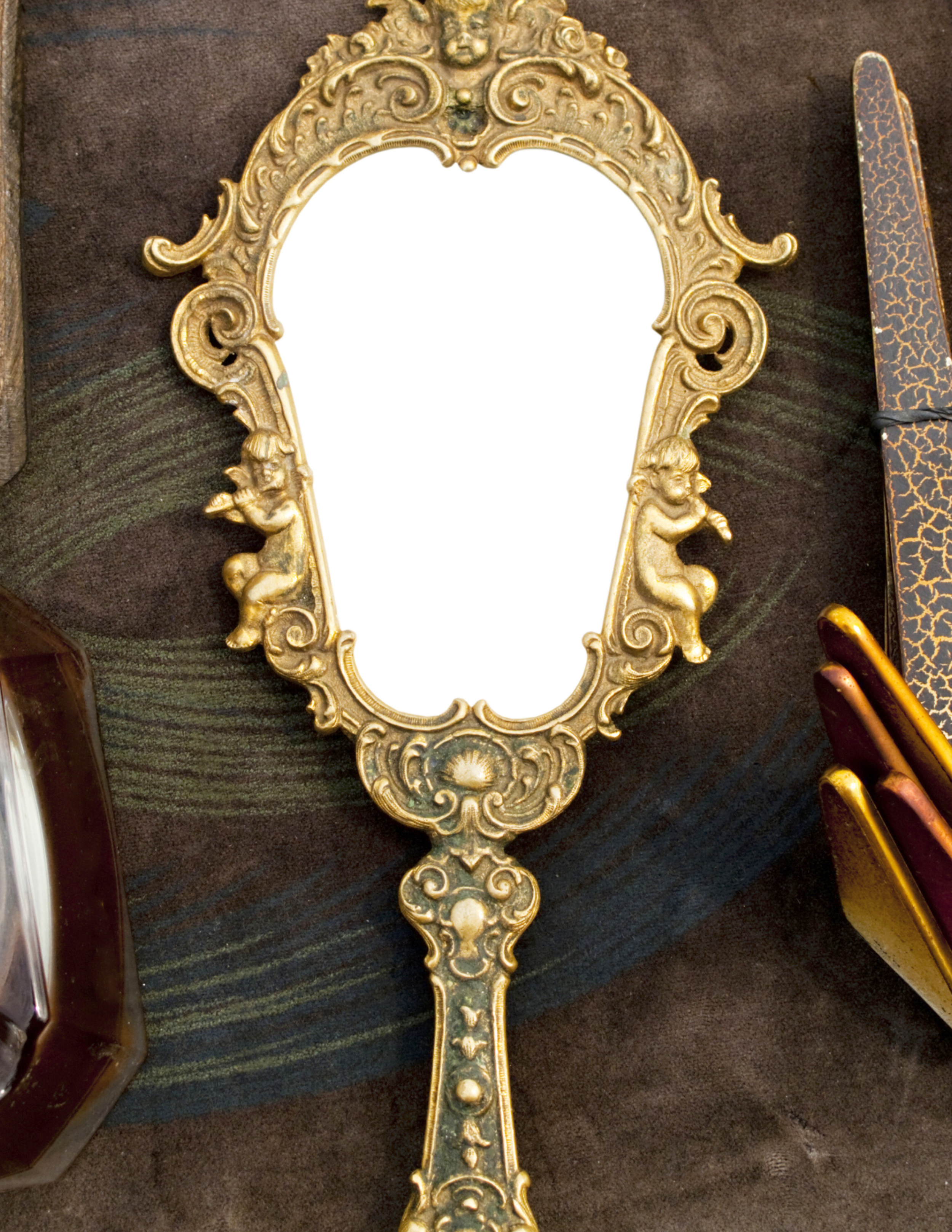 Ornate gold-colored mirror frame with cherub designs and decorative detailing, placed on dark surface next to a patterned object and a rod.