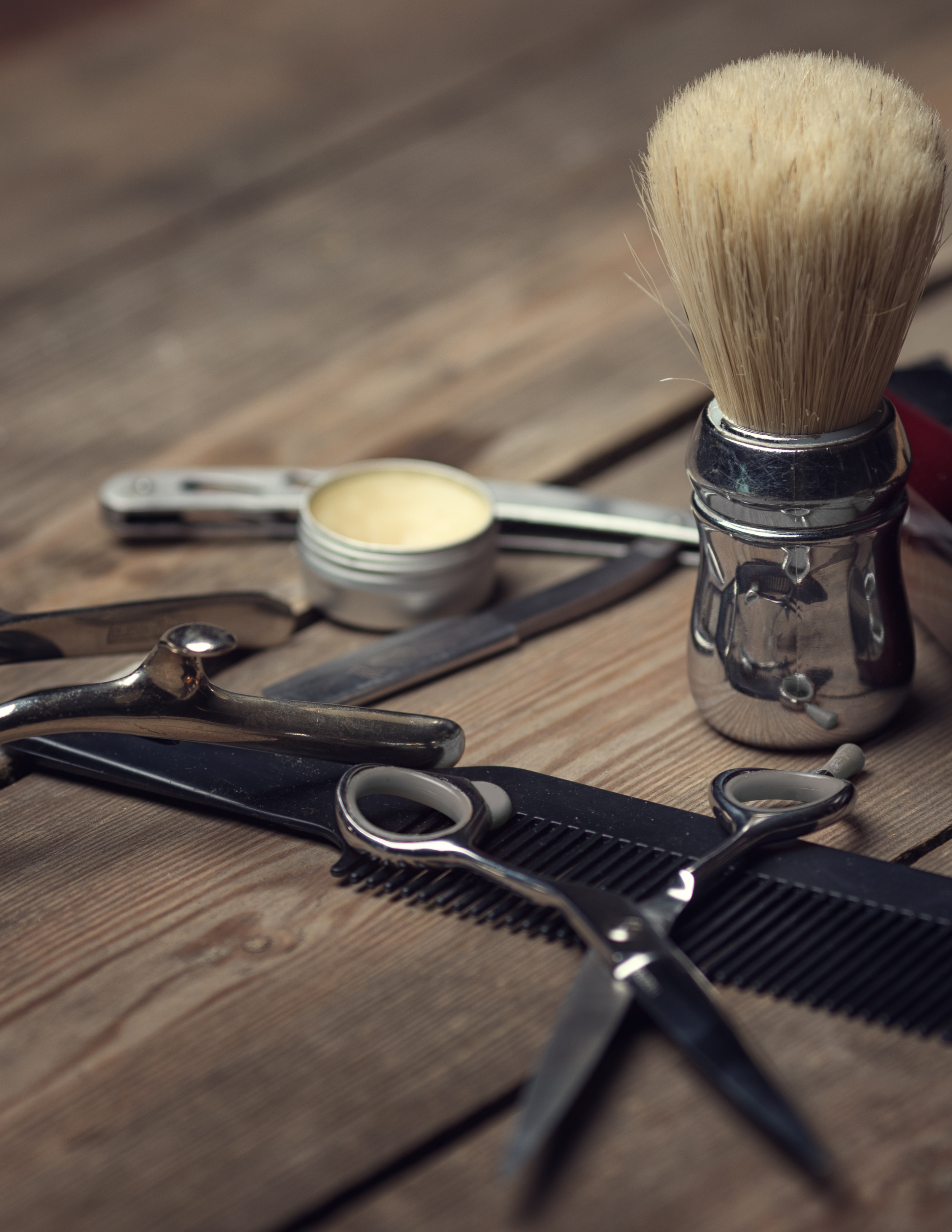 A collection of shaving tools including a shaving brush, safety razors, a comb, scissors, and a container of shaving cream on a wooden surface.