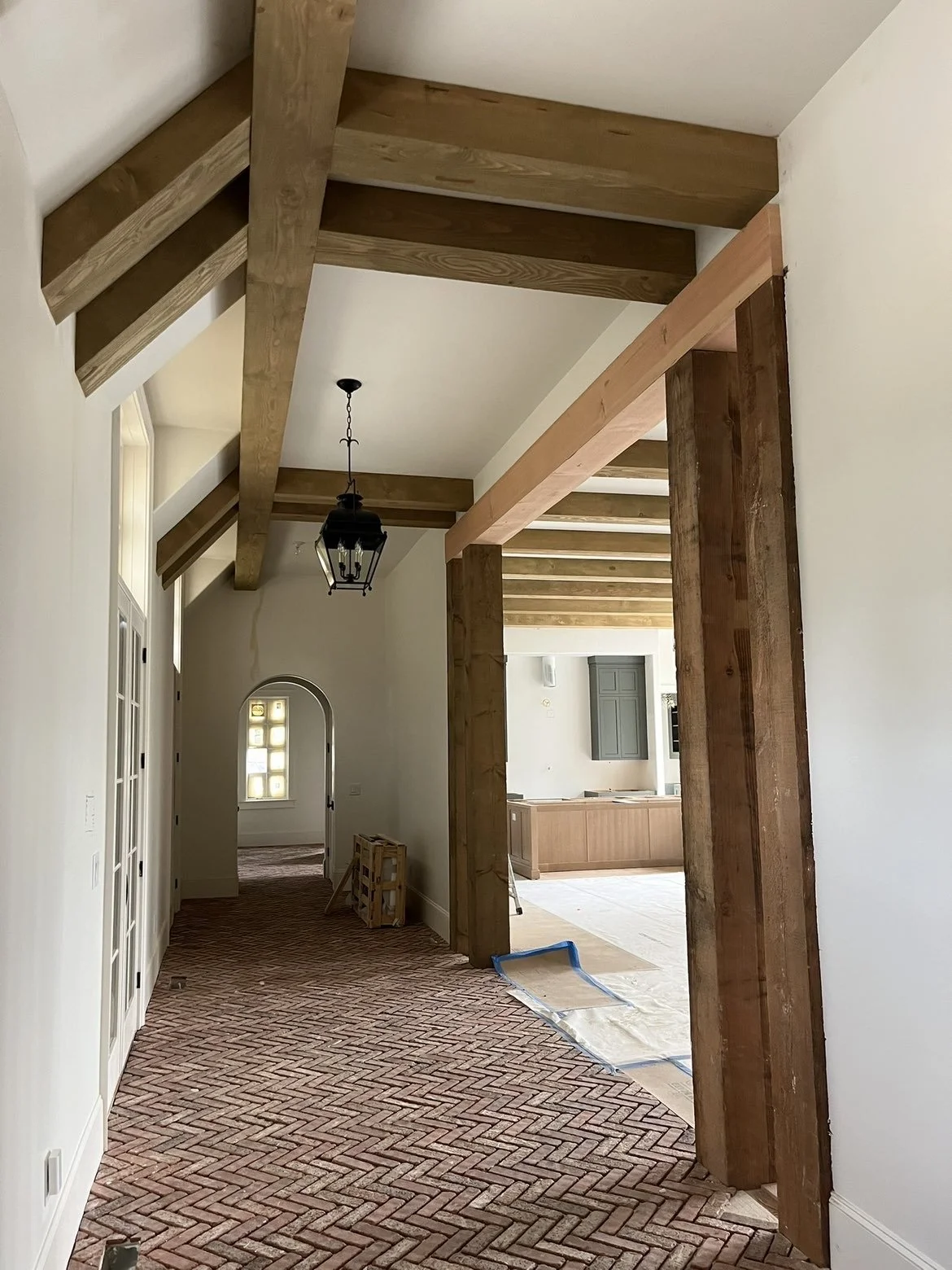 Interior of a house under renovation with exposed wooden beams, a hanging lantern, brick flooring, and an archway leading to another room with a window.
