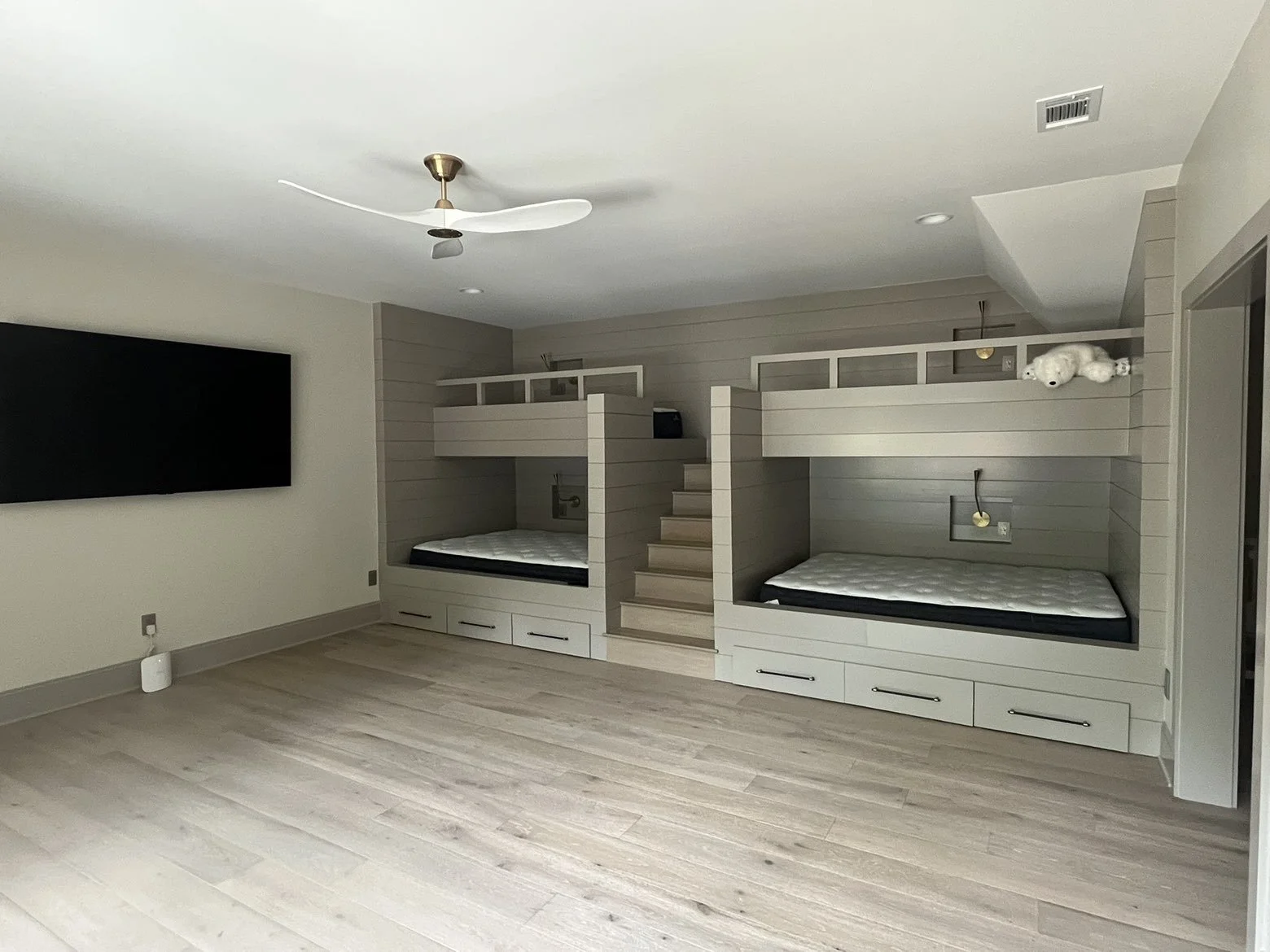 A bedroom with two built-in beds, each with mattresses, beneath a small loft area accessed by stairs. The room has a ceiling fan, a large wall-mounted television, and light-colored flooring.