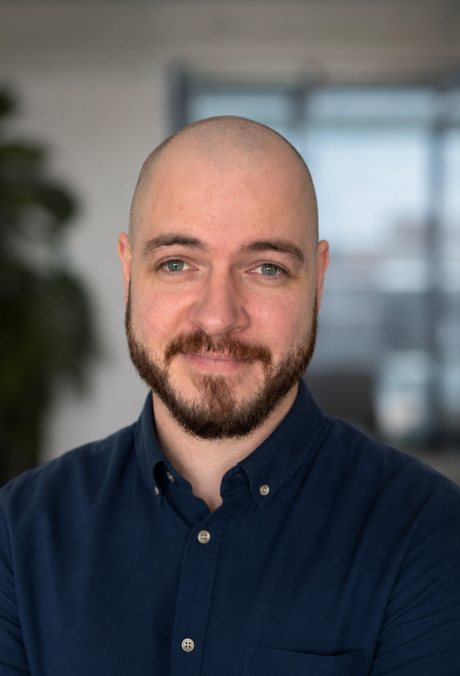 Headshot of a smiling man with a beard and shaved head, wearing a navy button-up shirt, standing indoors with blurred background.