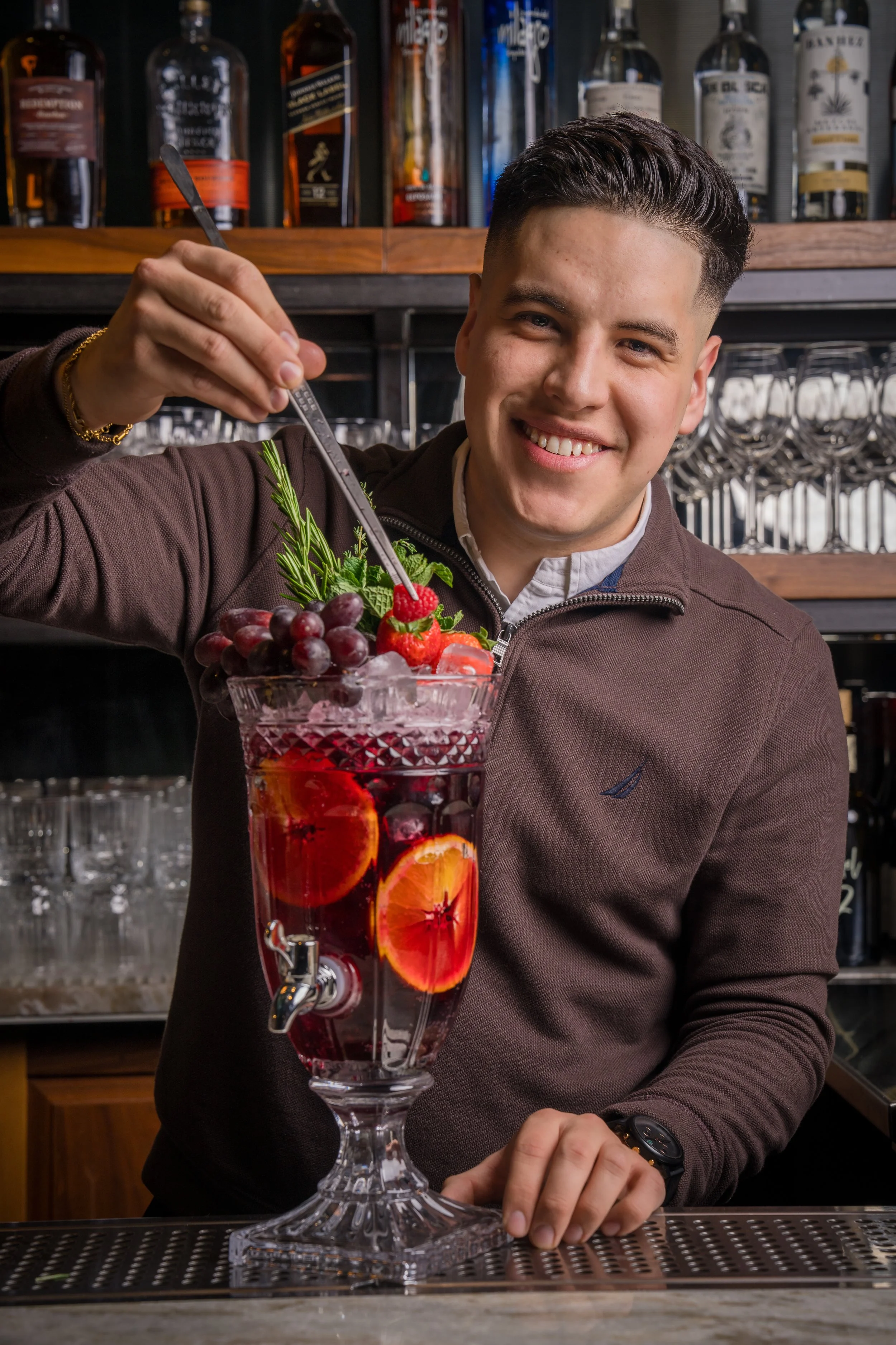 A bartender smiling and holding a metal cocktail pick over a large, decorative glass filled with red drink, fruit slices, strawberries, grapes, and mint, in a bar setting with bottles and glassware in the background.