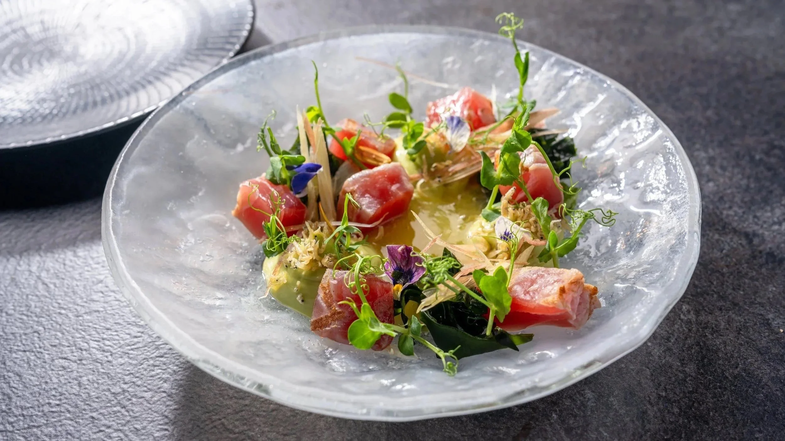 A gourmet salad with raw fish, microgreens, edible flowers, and a light vinaigrette dressing served in a textured glass bowl.