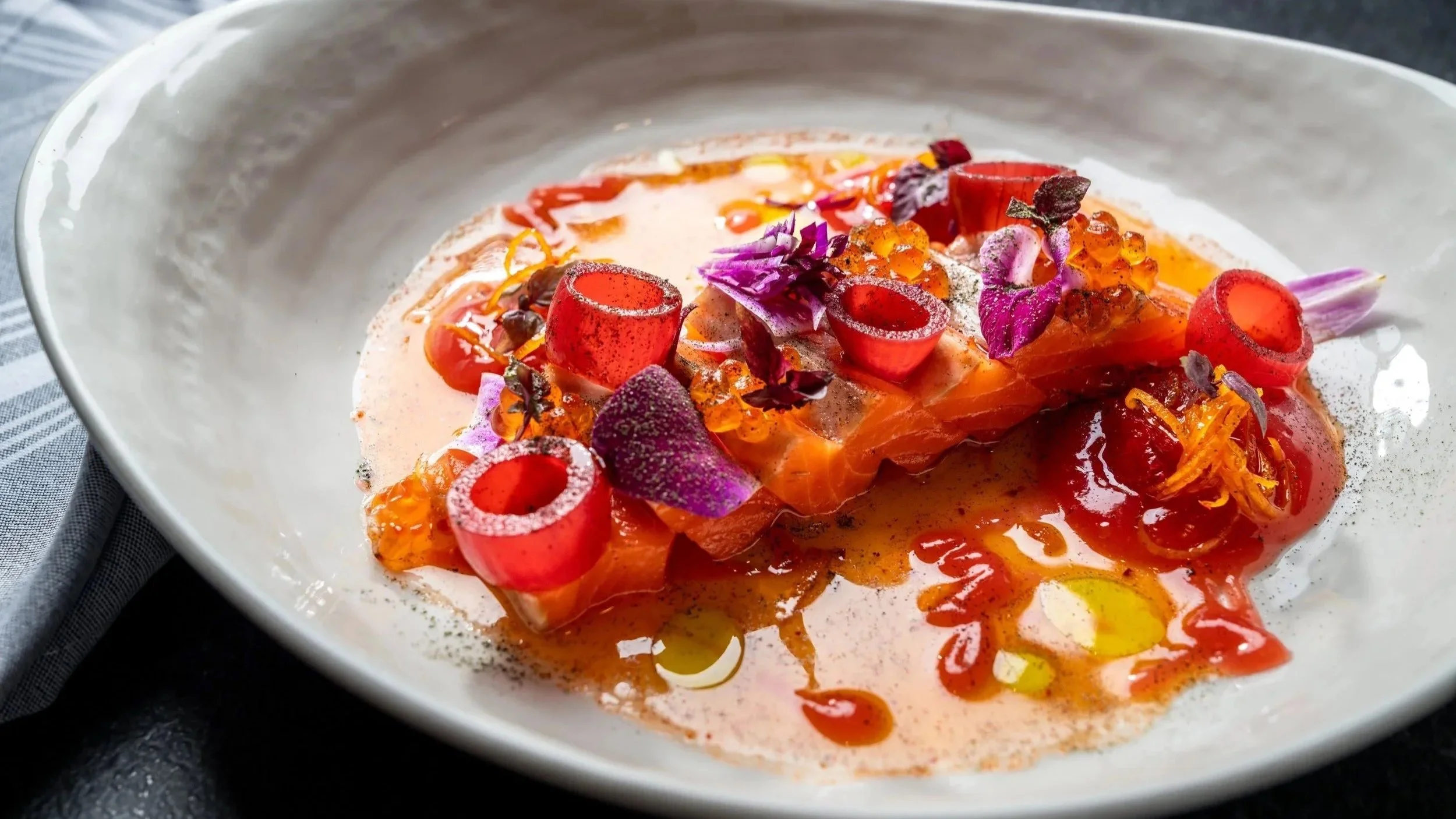 A plated dish of smoked salmon topped with edible flowers, roe, and herbs, garnished with a sauce.