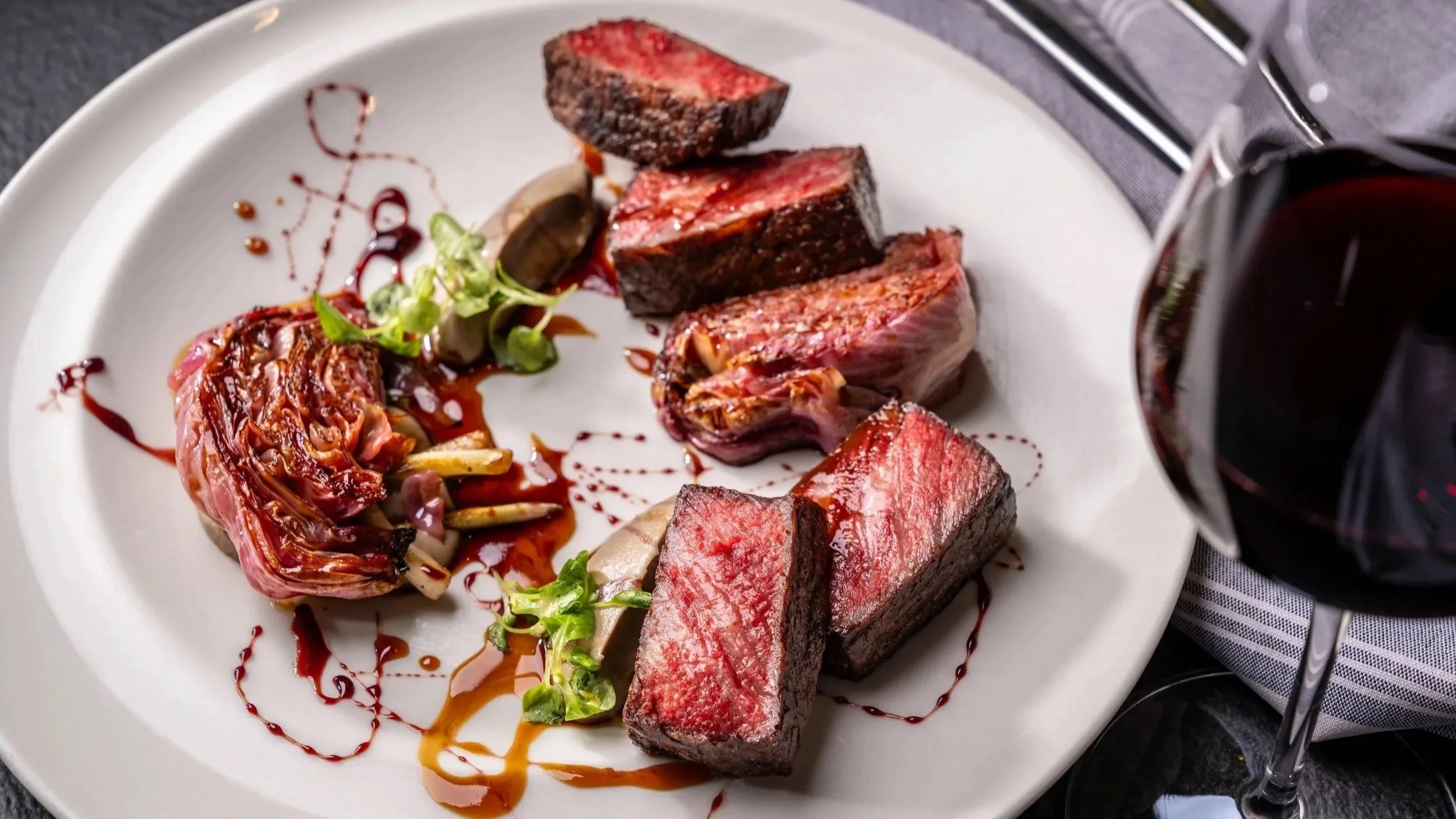 A white plate with slices of medium-rare steak, roasted vegetables, and a garnish, with a glass of red wine on the side.