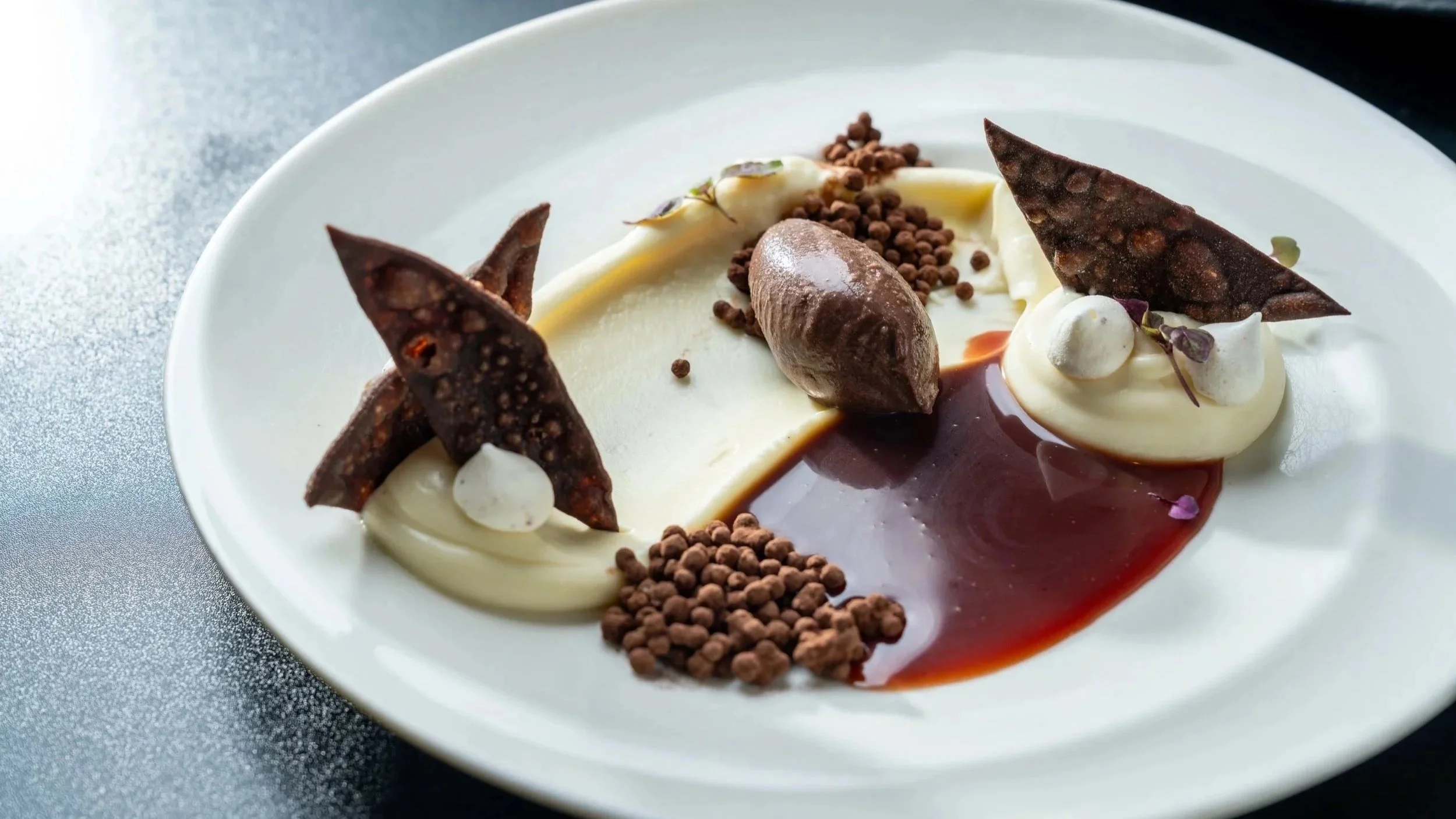 A plated dessert with chocolate foam, chocolate shards, chocolate pearls, and a decorative sauce on a white plate.