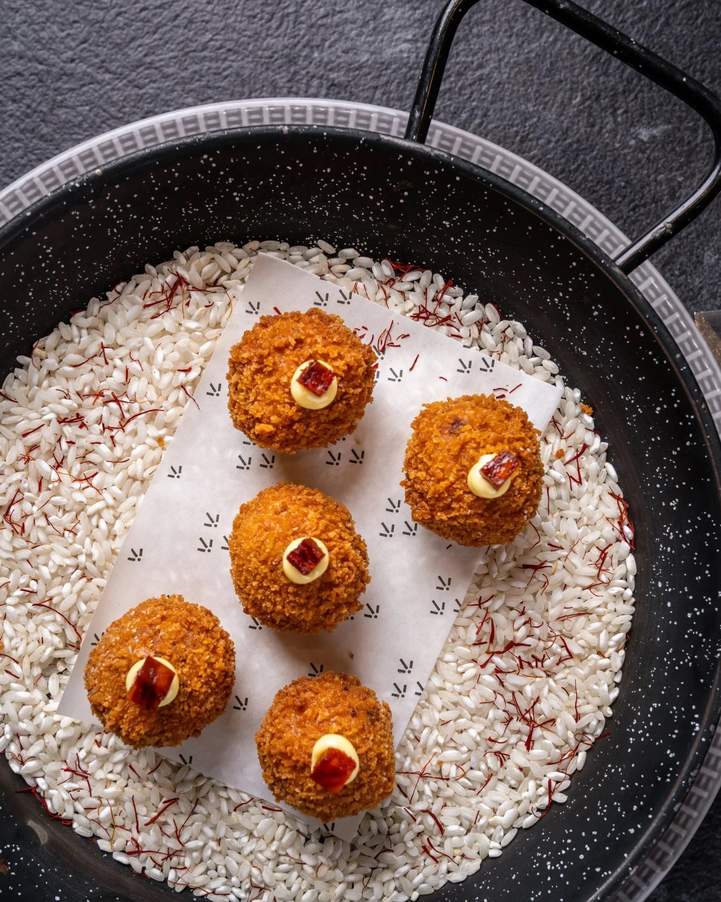 Deep-fried food balls topped with small dollops of white sauce and a small piece of red garnish, placed on a sheet of white paper inside a black metal tray filled with small white rice grains and red saffron strands, on a dark grey surface.