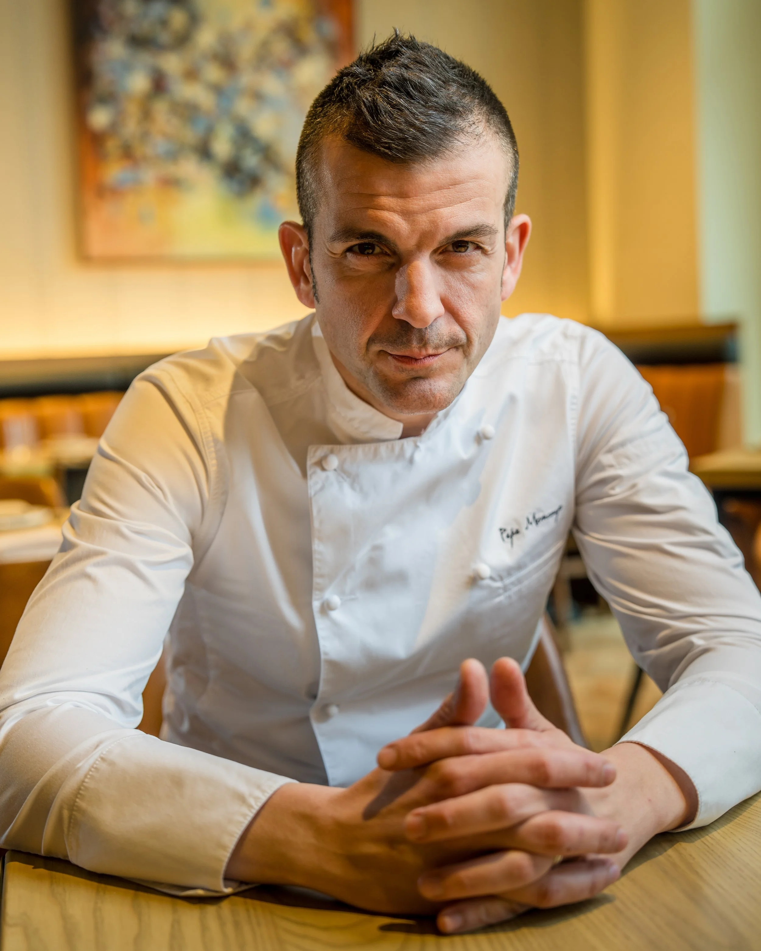 Close-up of a male chef with short dark hair, wearing a white chef's coat, sitting at a wooden table with his hands clasped together, in a restaurant or cafe setting.