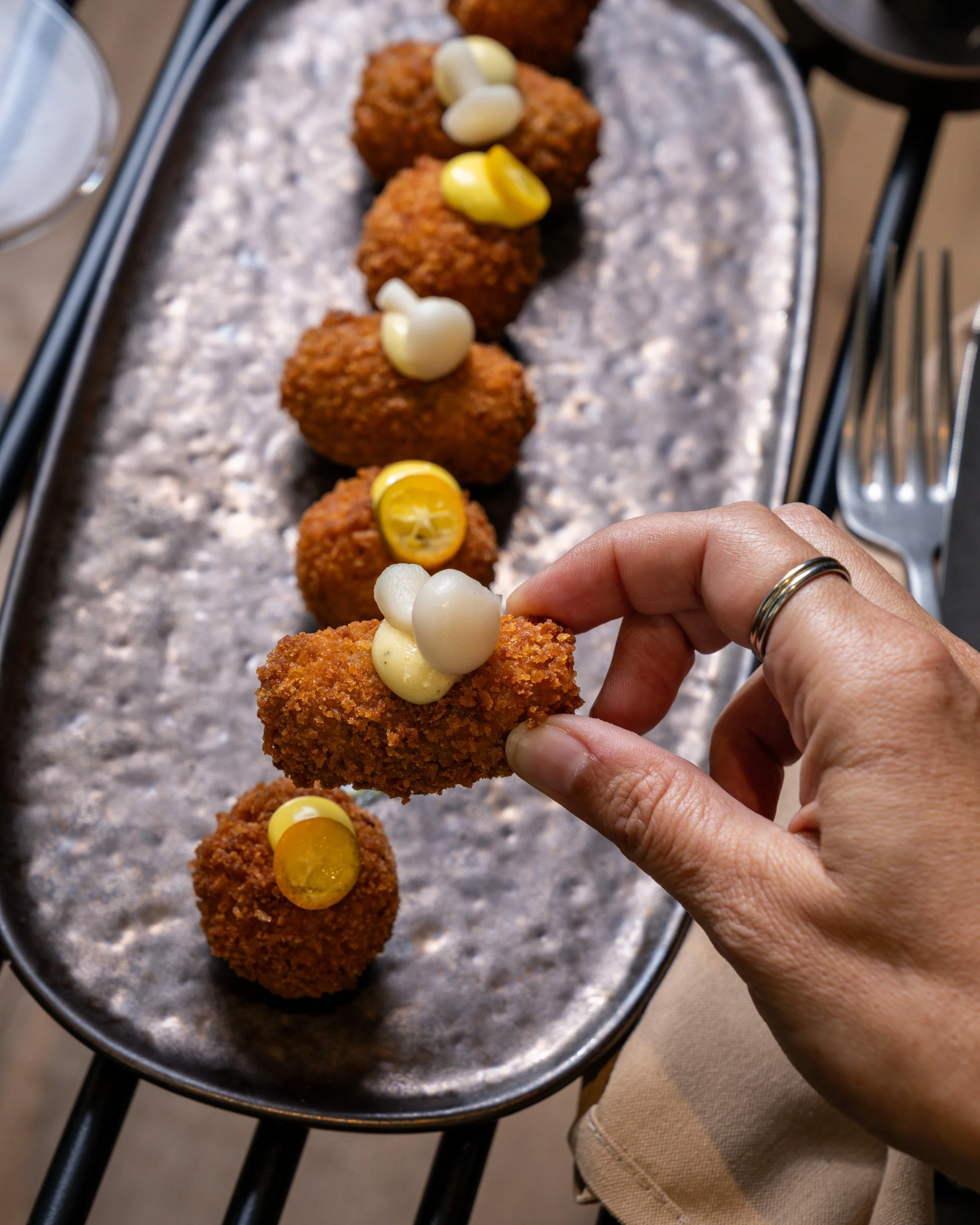 Hand holding a fried food item topped with garlic and lemon slices, with more similar fried items lined up on a metallic tray in the background.