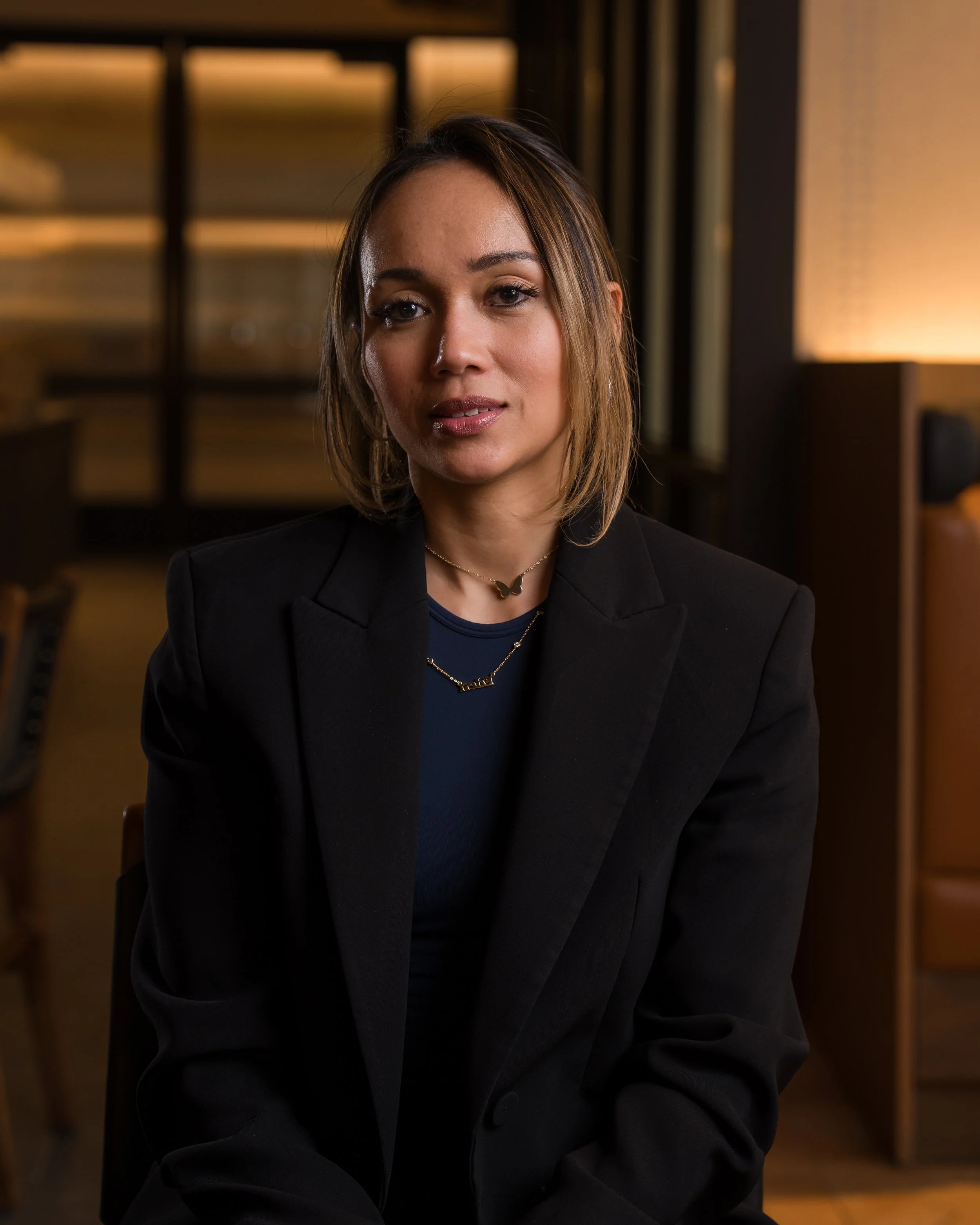 A young woman with shoulder-length brown hair and makeup in a dimly lit room with a background of shelves and warm lighting, wearing a black blazer, necklace, and blue top.