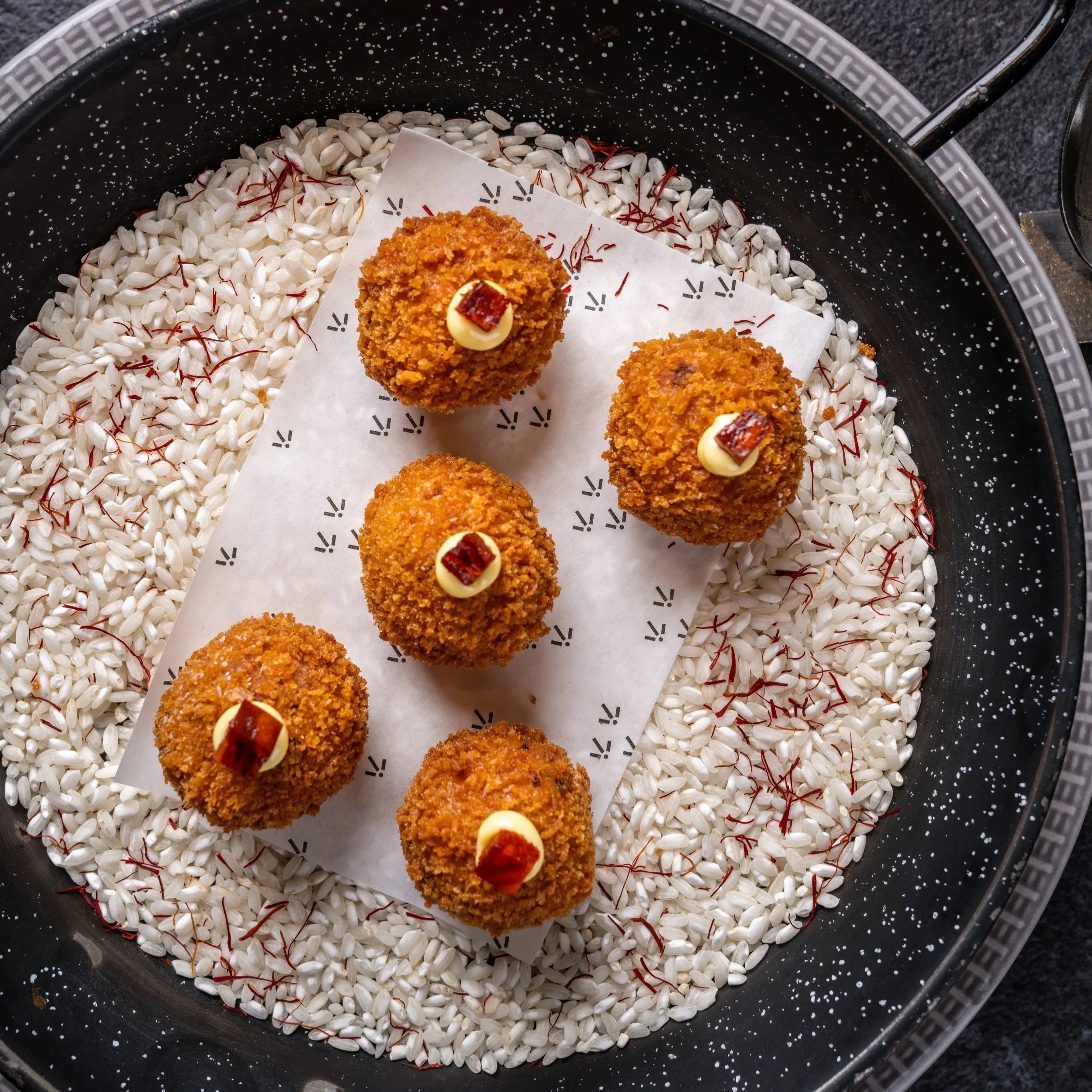 Close-up of breaded, fried food balls topped with small dollops of white sauce and bits of red dried fruit, arranged on white paper inside a black, speckled pan filled with white grains.