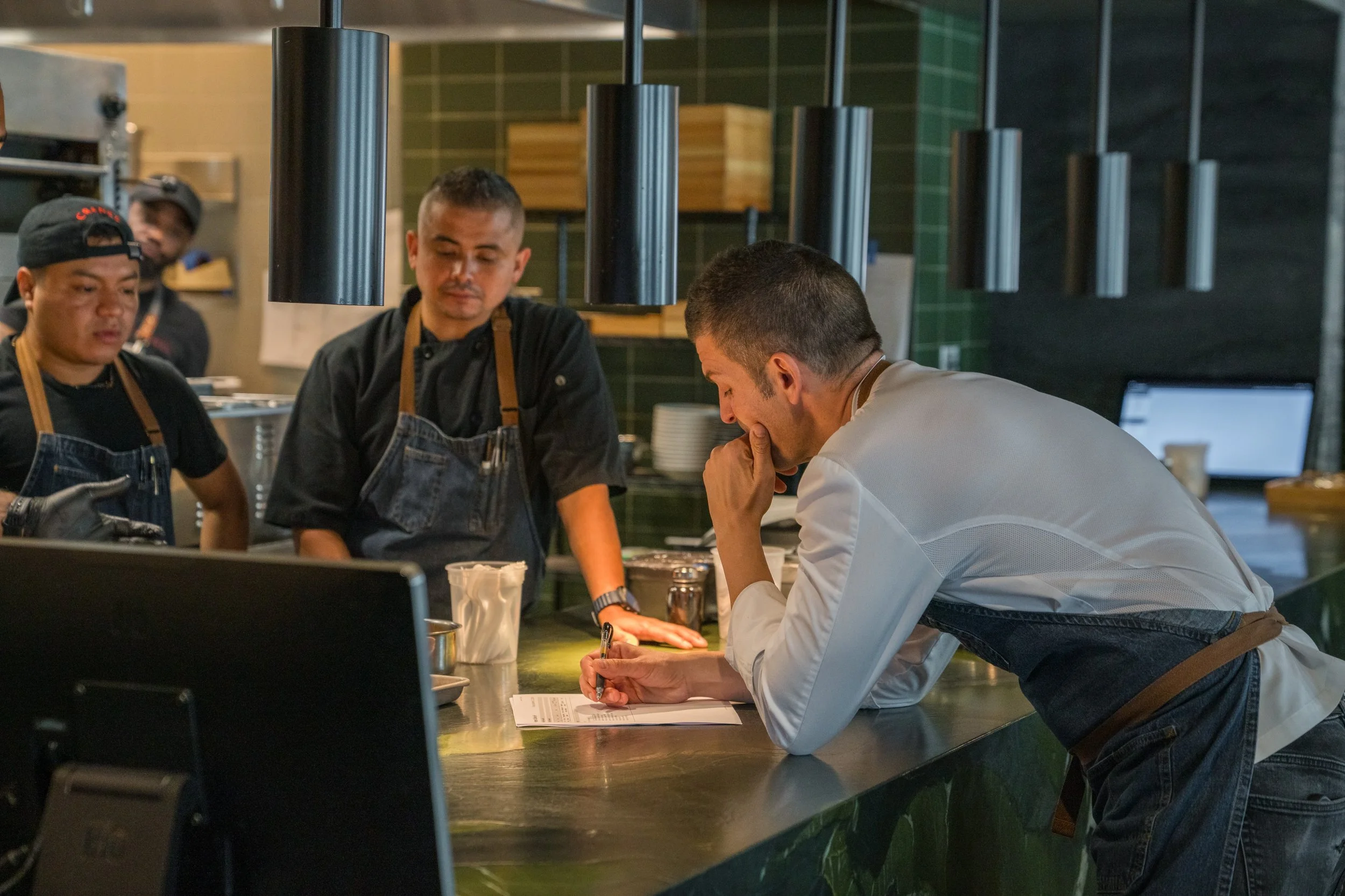 Three chefs and a customer in a restaurant kitchen, one chef is writing notes while the others observe.