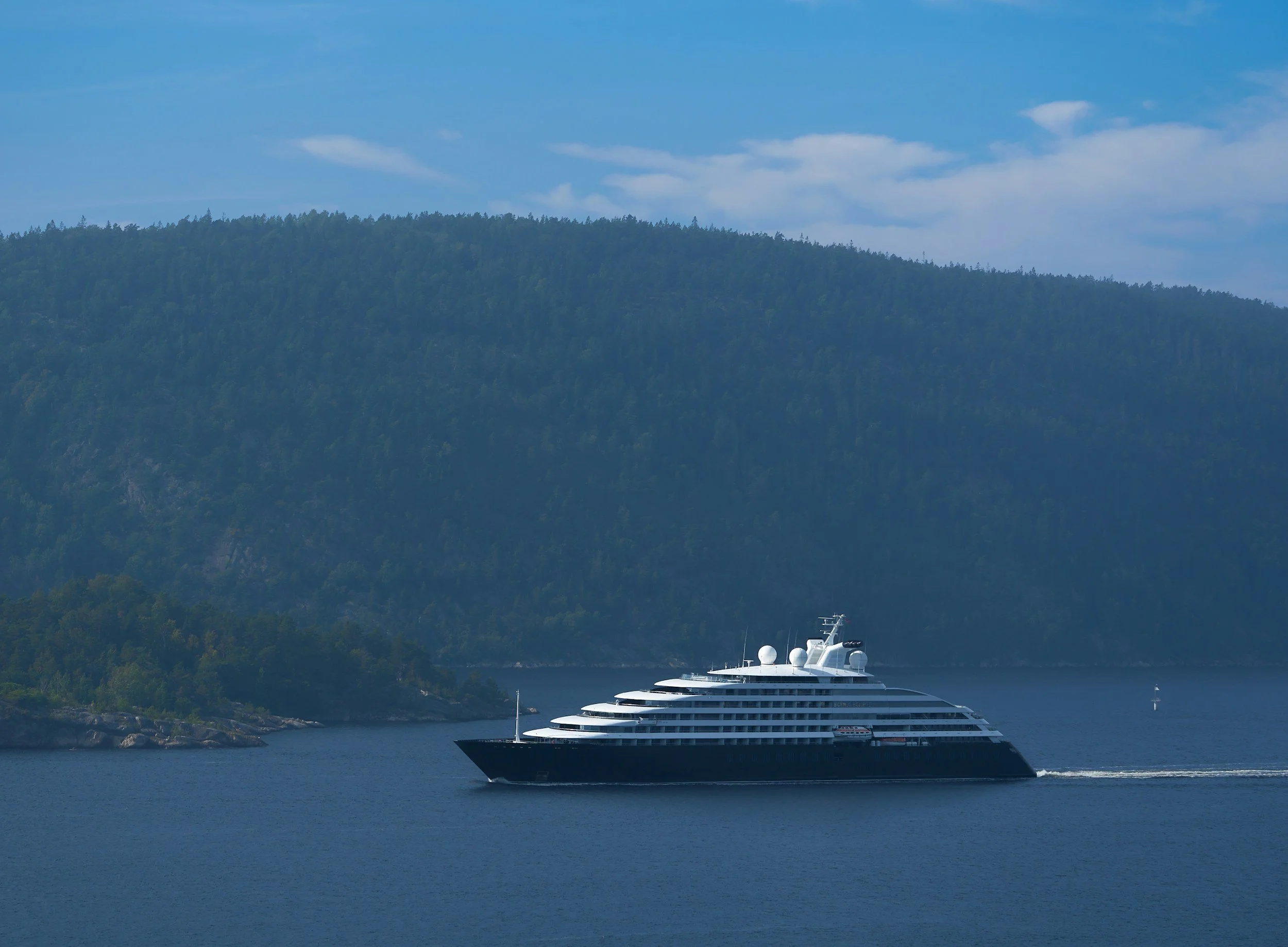 A large white cruise ship sailing in a body of water with a forested hill in the background under a blue sky with some clouds.