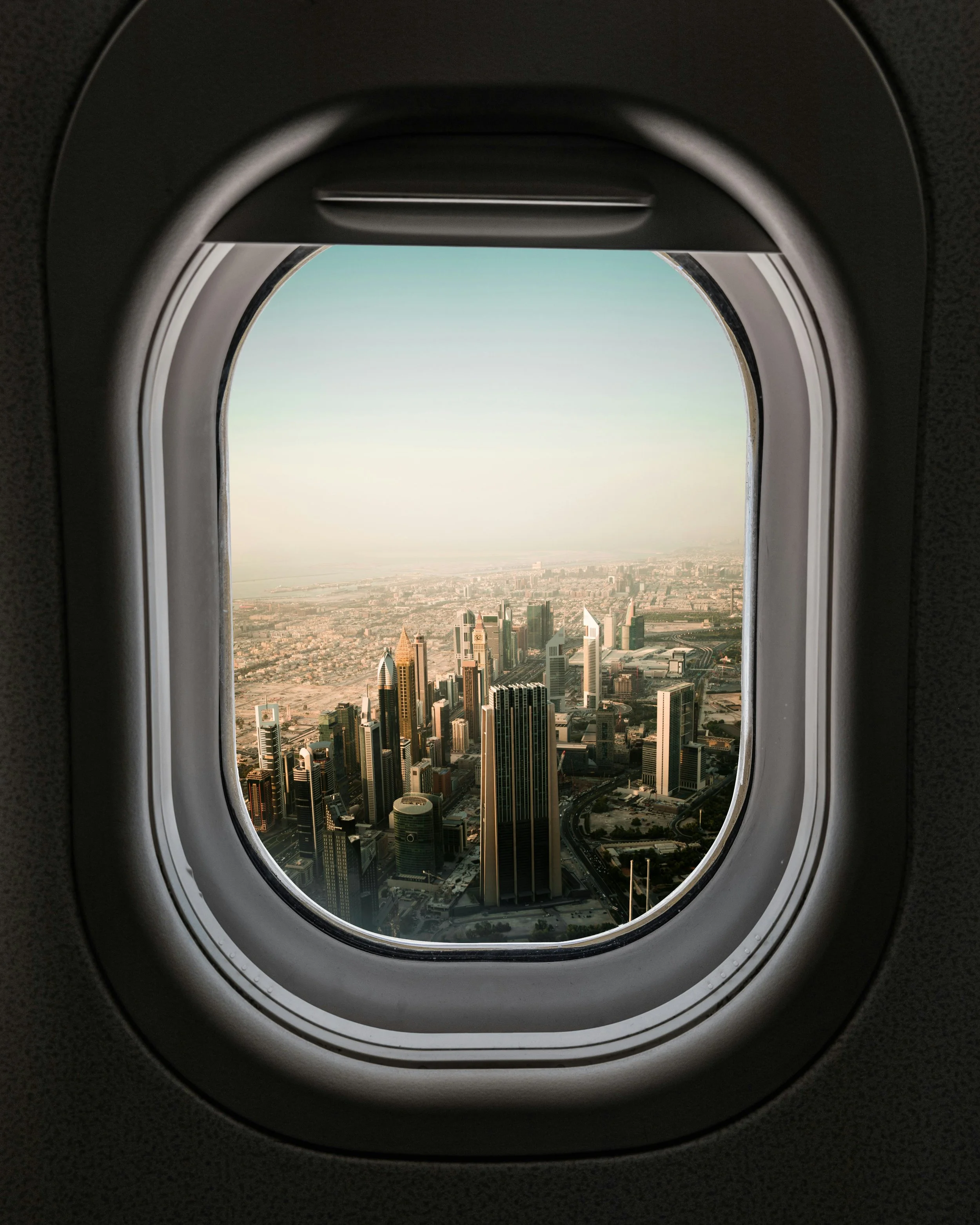 View of a city skyline through an airplane window with a rounded rectangular shape, showing tall skyscrapers against a clear sky.