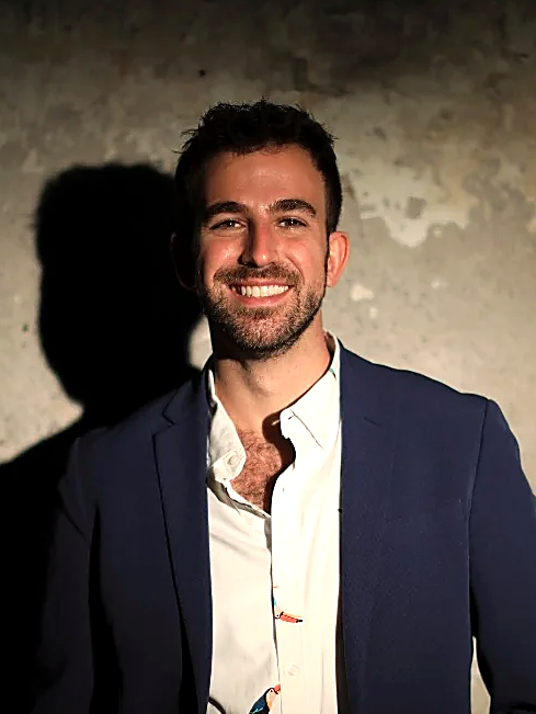 A smiling man with dark hair and a beard wearing a white shirt and a dark blazer, standing against a textured gray wall.