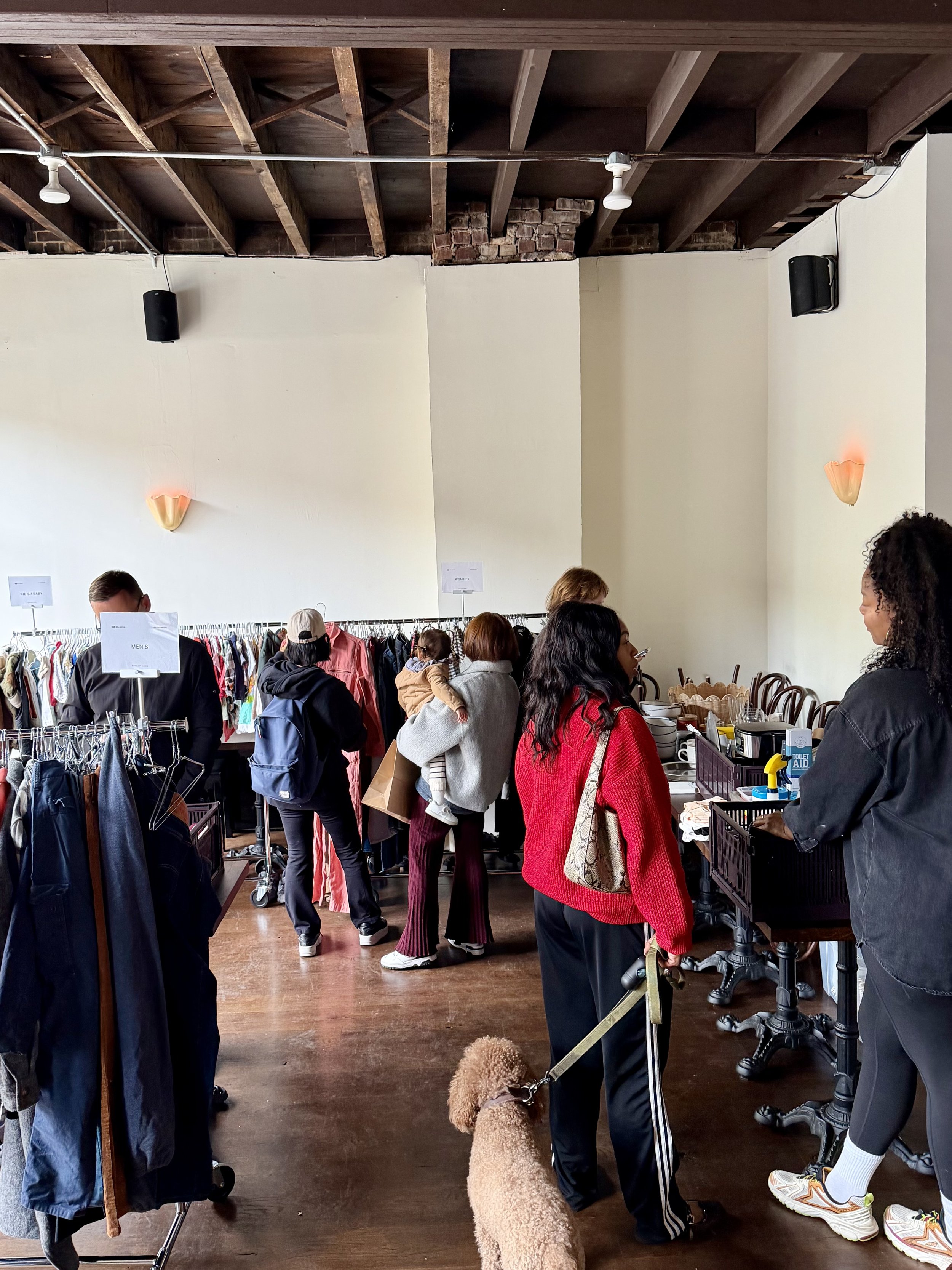 People shopping at a clothing swap at Cicchetti with exposed ceiling beams and bright lighting.