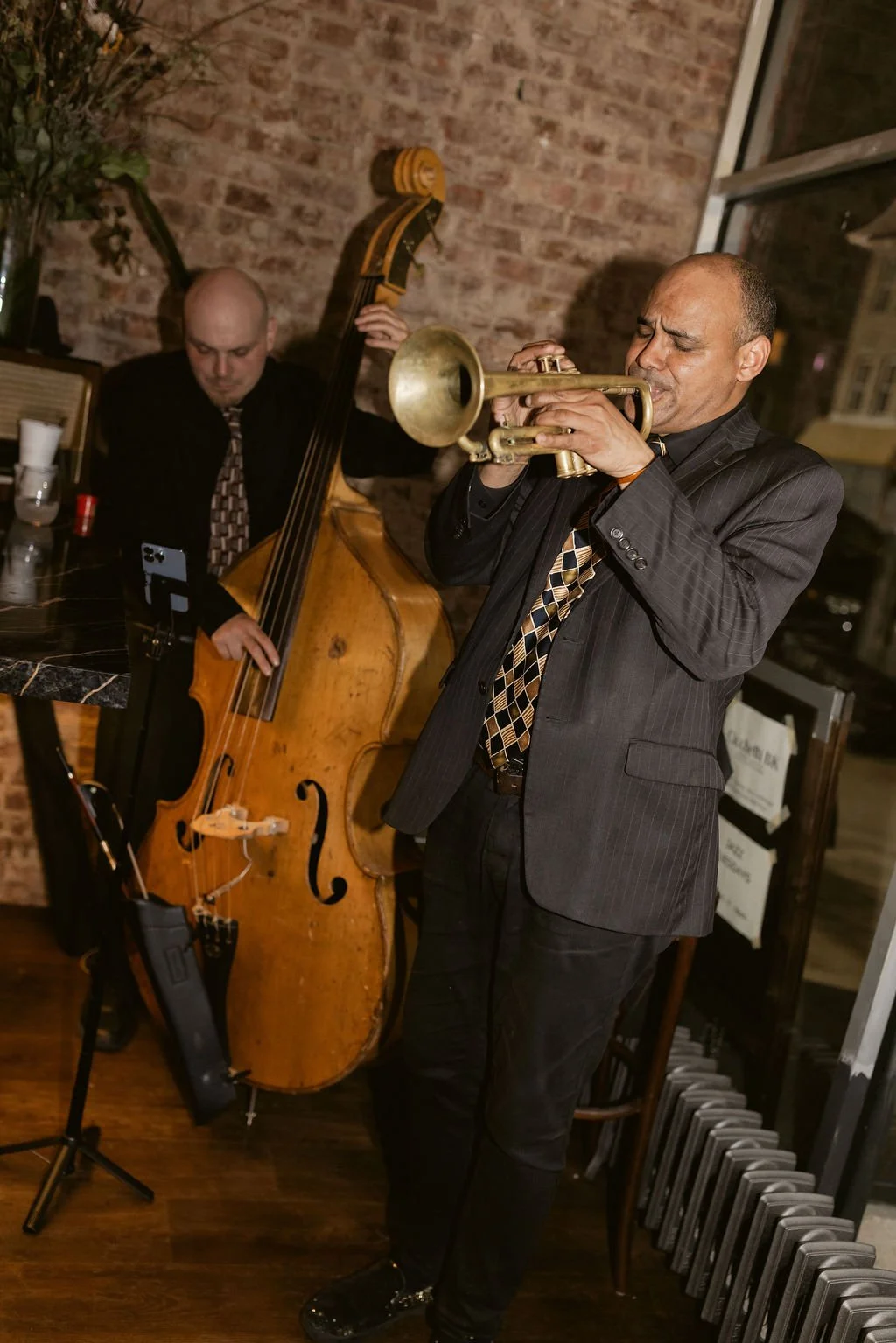 Two musicians performing indoors; one playing a trumpet and the other playing a double bass, with a brick wall background.