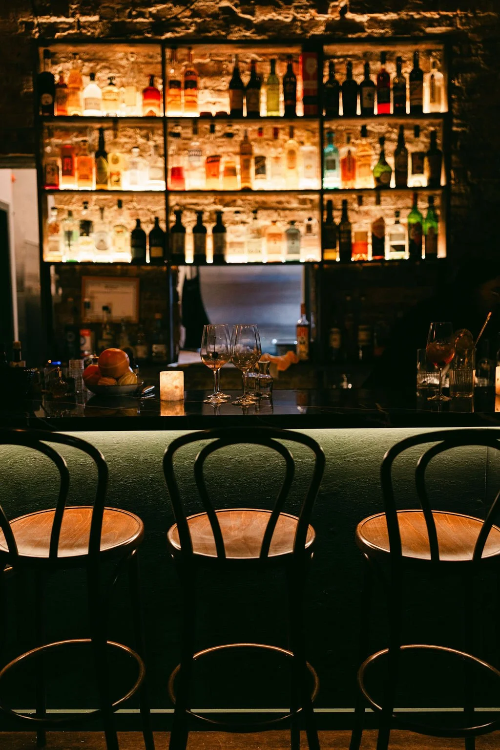 Bar with bar stools, bottles of alcohol on backlit shelves, wine glasses on the counter, and a candle, in a dimly lit setting.