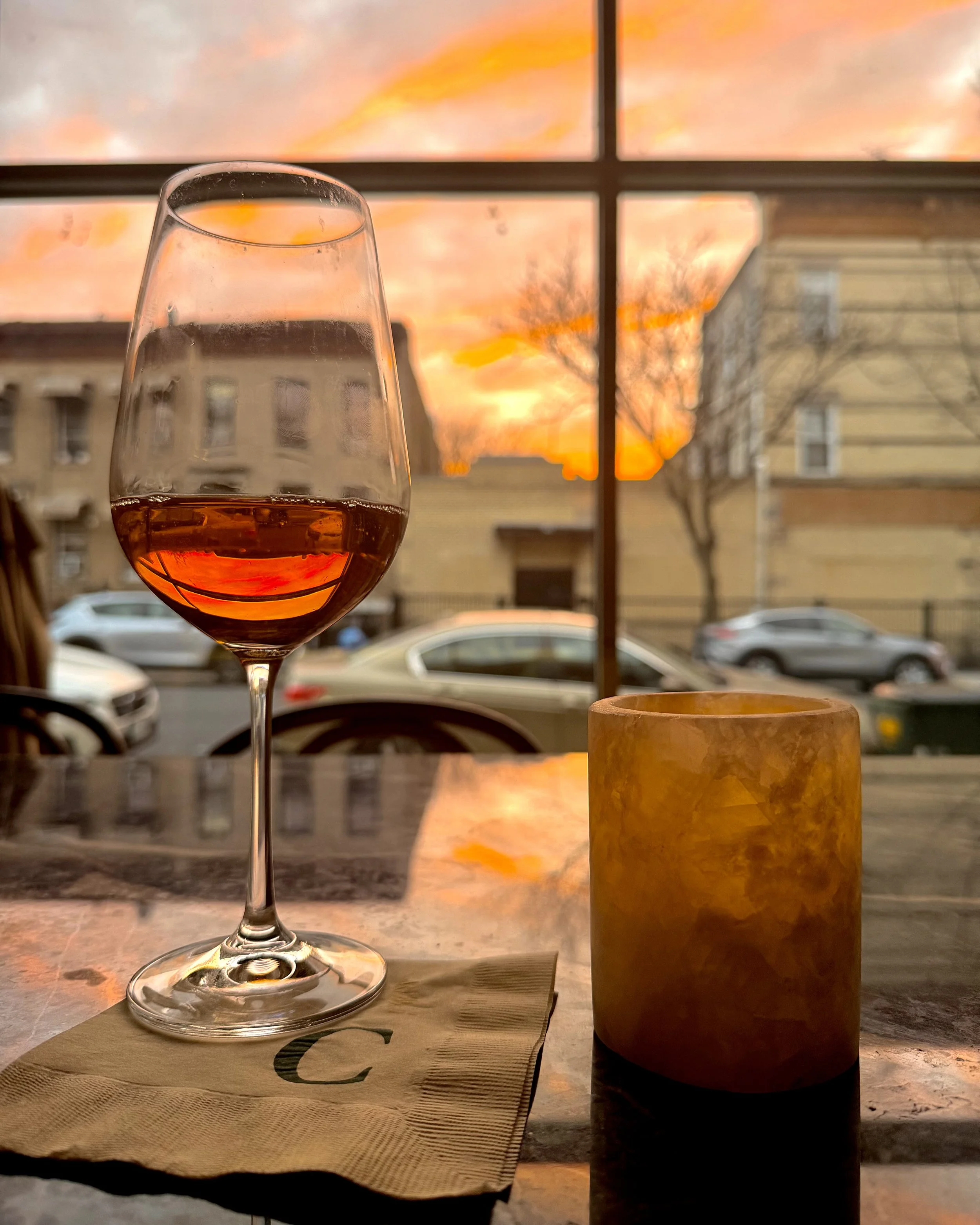 A glass of rose wine on a table with a sun setting outside through a window, with buildings and cars in the background.