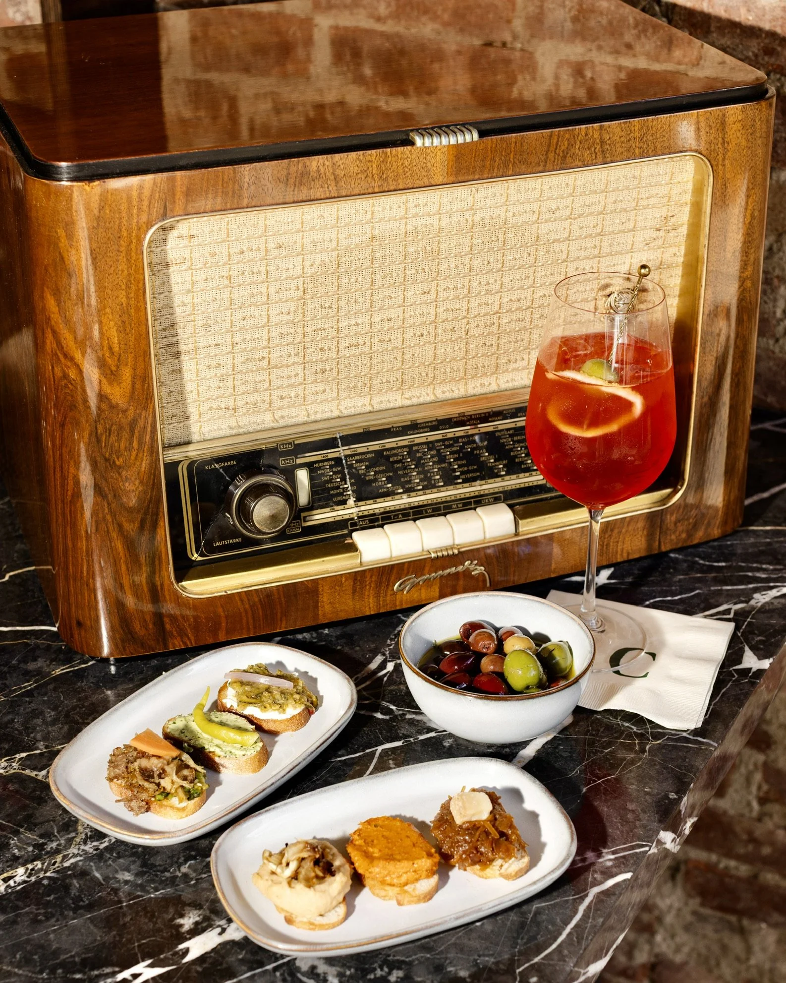 A vintage wooden radio with a food and drink setup in front of it, including an assortment of small appetizers on white dishes, a bowl of mixed olives, and a glass of red cocktail garnished with fruit.