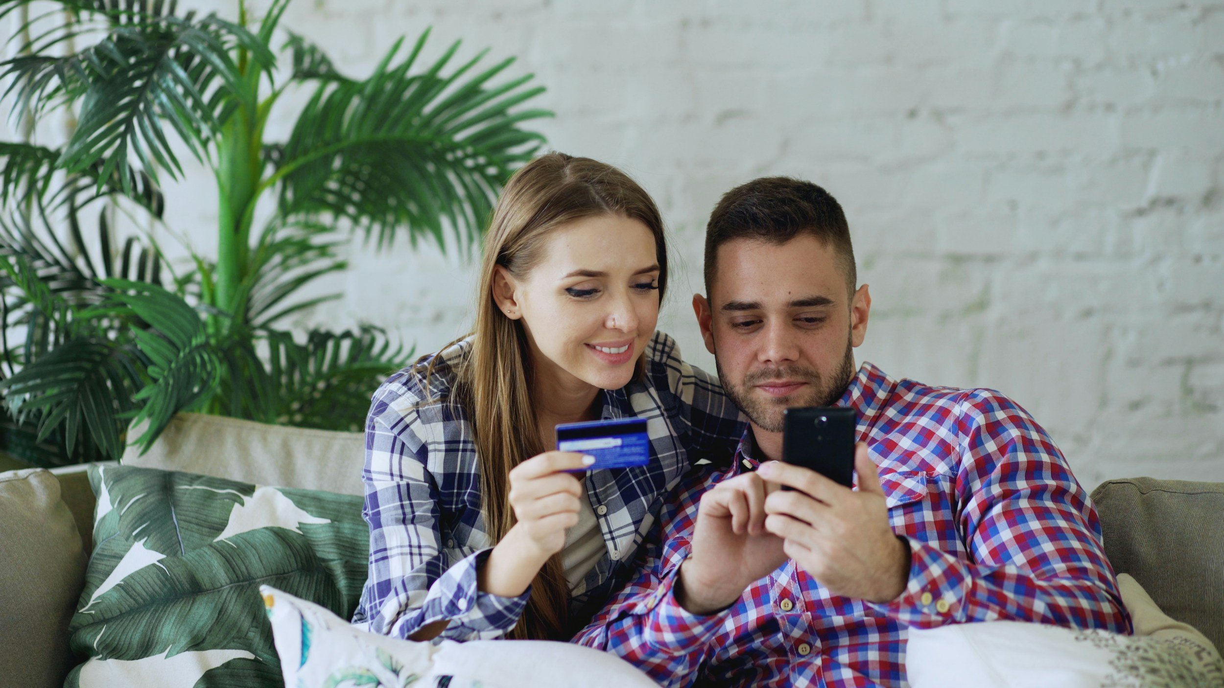 A young woman and a young man sitting on a beige couch, looking at a smartphone together. The woman holds a credit card in her hand. They are in a bright room with a white brick wall and green plants.