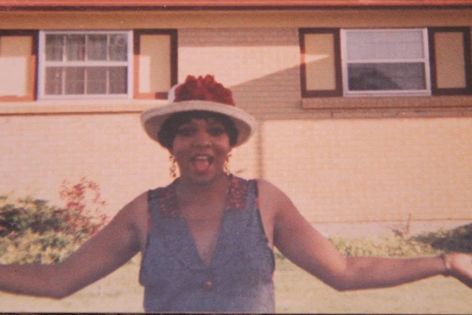 A woman wearing a dress with decorative shoulder details and a wide-brimmed hat with red flowers, smiling with arms outstretched in front of a house with brick walls and multiple windows.
