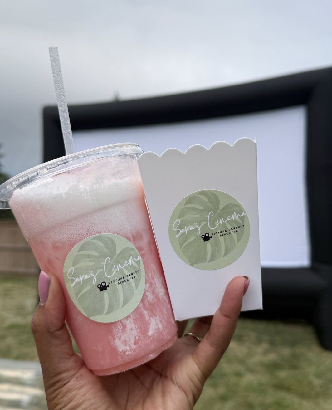 A person holding a pink frozen beverage in a clear plastic cup with a straw, and a white box with a green logo that says 'Sara's Cinema' with a leaf design, outdoors with a blurry outdoor movie screen in the background.
