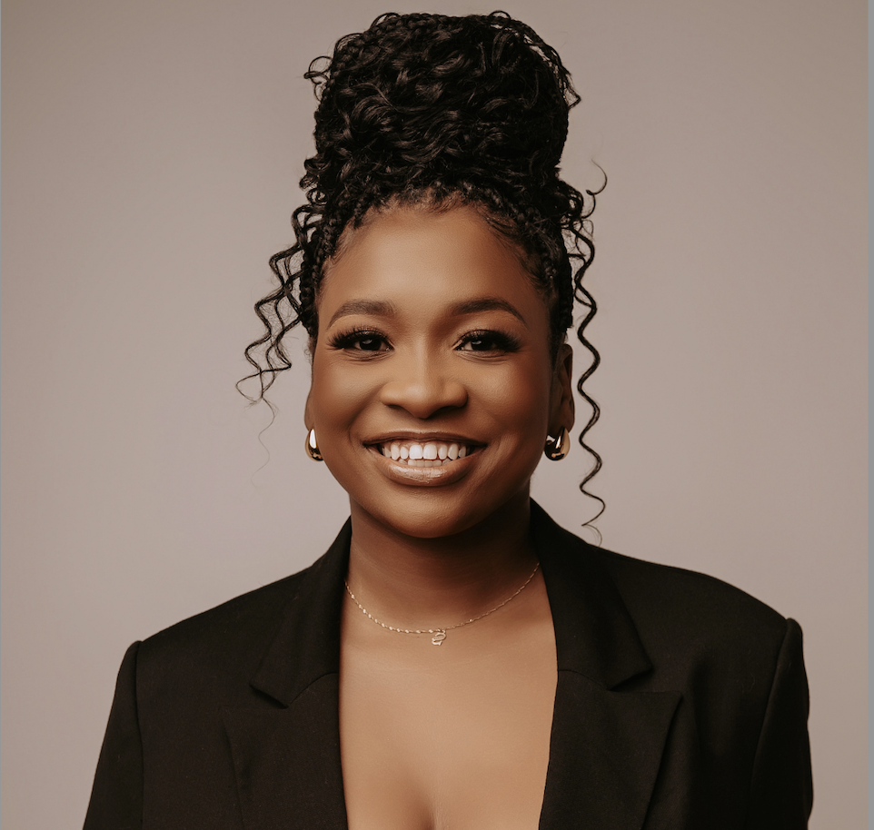 Portrait of a smiling Black woman with curly hair styled in an updo, wearing a black blazer, gold earrings, and a delicate gold necklace, against a plain neutral background.